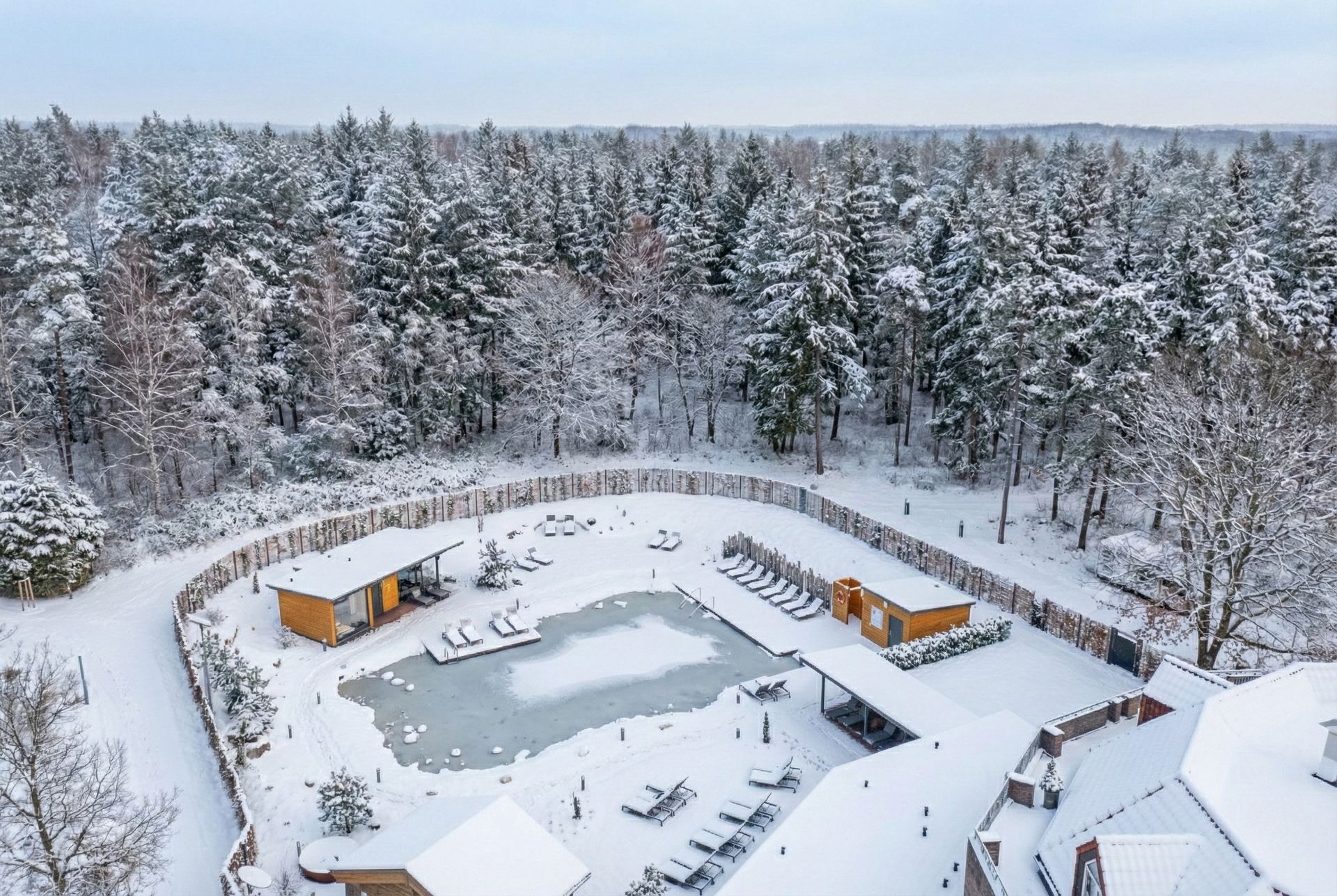 Luftaufnahme eines kleinen, zugefrorenen Schwimmbeckens, umgeben von schneebedecktem Gelände und einem dichten Wald im Winter.