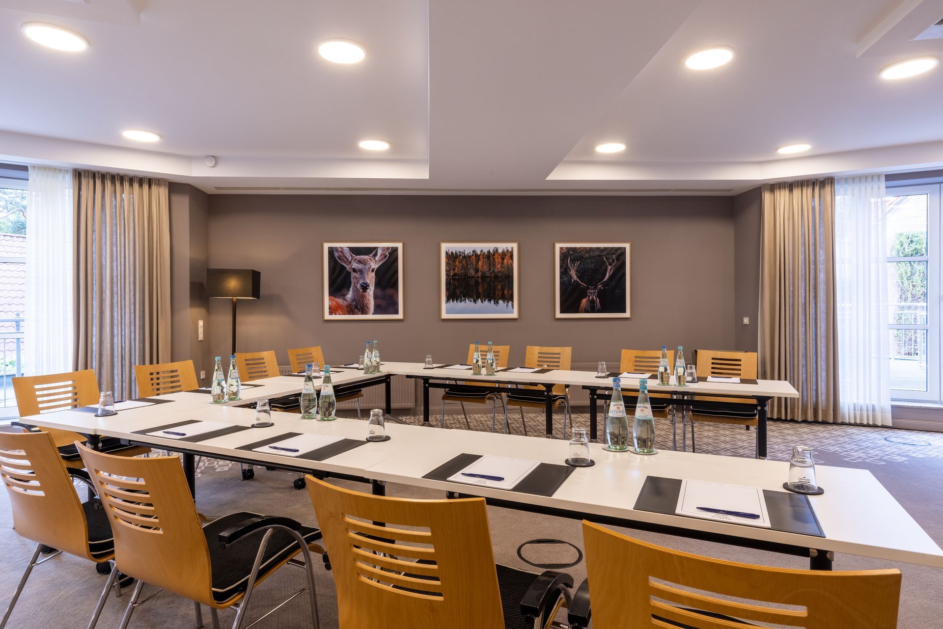 A modern conference room decorated in neutral tones, featuring a U-shaped table arrangement and wooden chairs aligned with three framed photos.