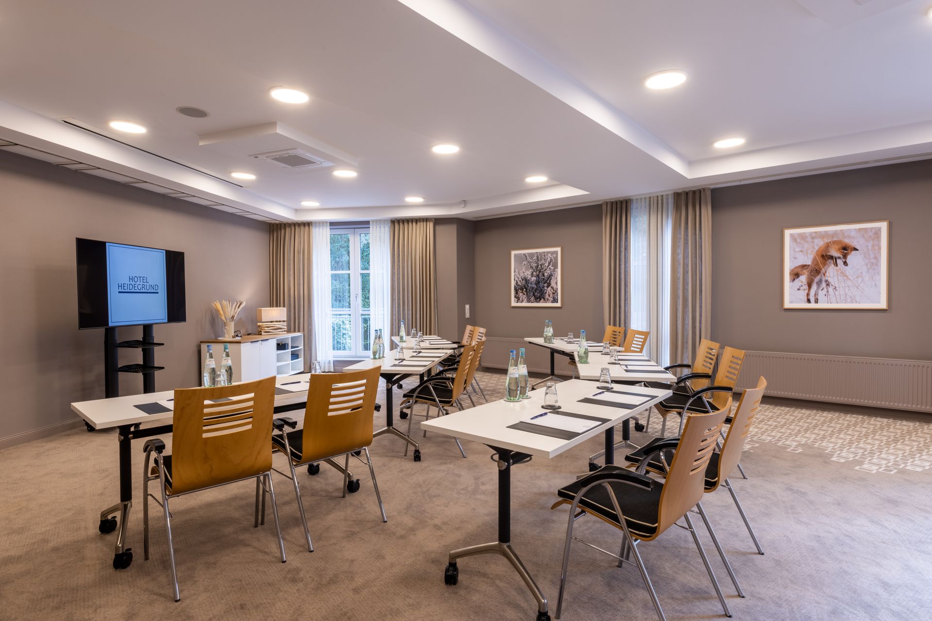 Ein hell erleuchteter Konferenzraum mit Reihen von Tischen und Holzstühlen, die auf einen großen, an der Wand montierten Monitor ausgerichtet sind.
