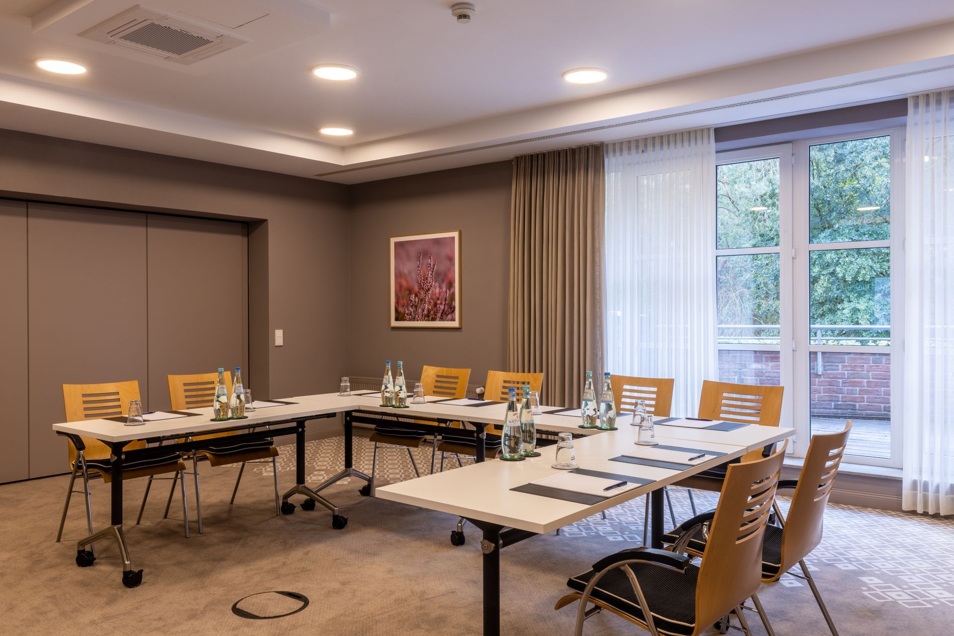 A meeting room with white tables arranged in a U-shape and light-colored wooden chairs on a beige carpet.