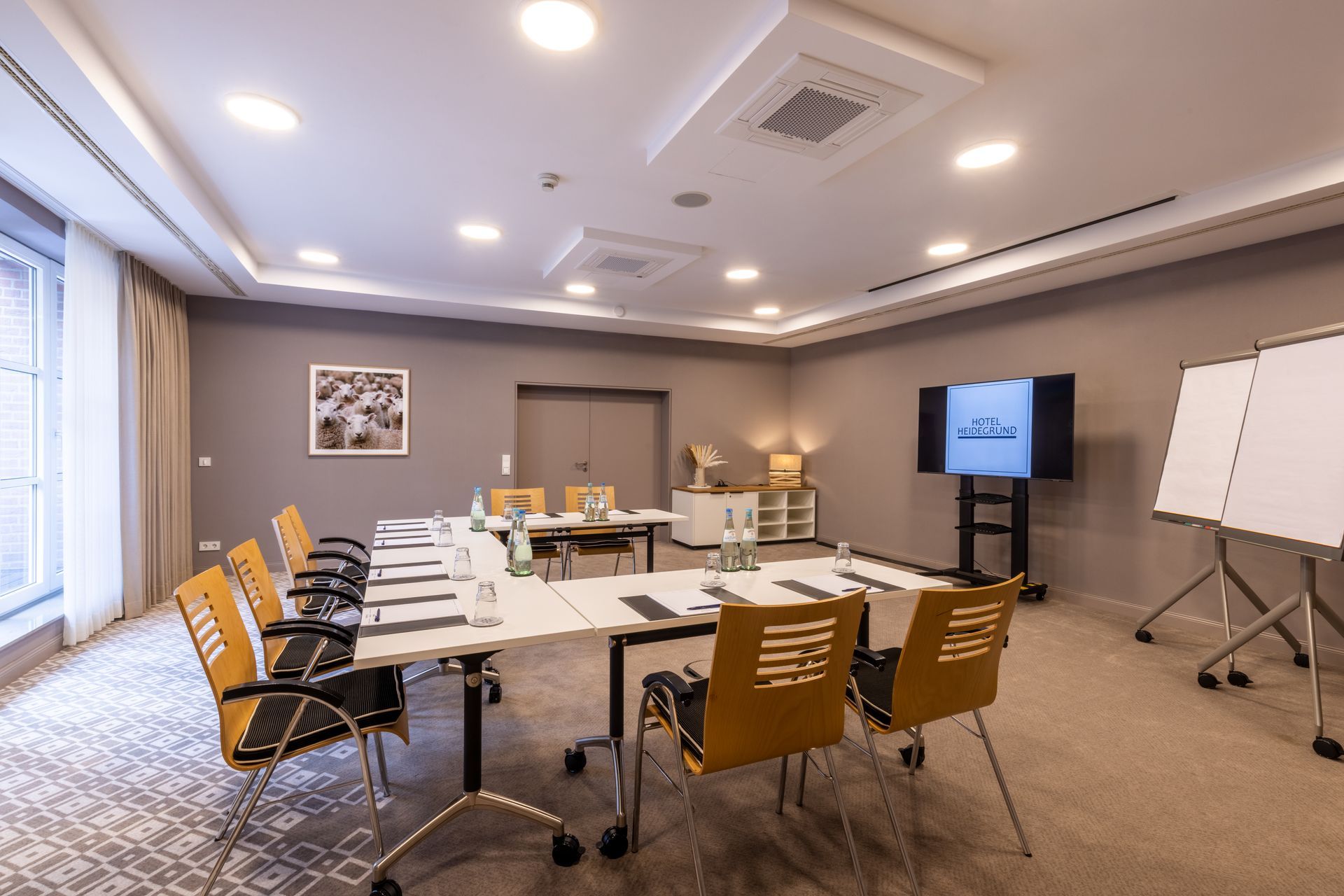 Ein moderner, in neutralen Tönen gehaltener Konferenzraum mit U-förmiger Tischanordnung, orangefarbenen Stühlen, einem Fernseher und Whiteboards.