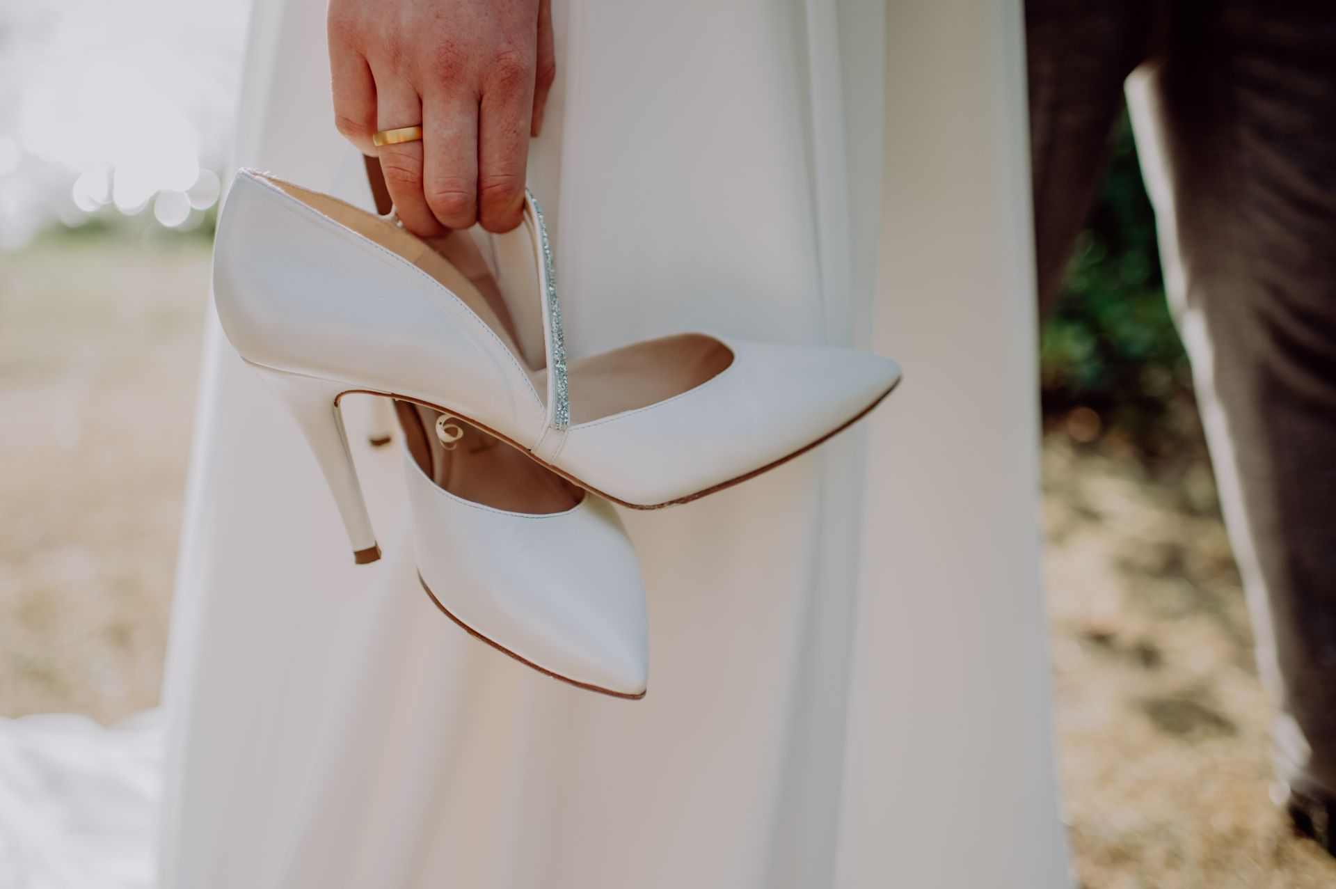 Eine Person im Brautkleid hält ein Paar weiße, spitz zulaufende High Heels am Absatz.