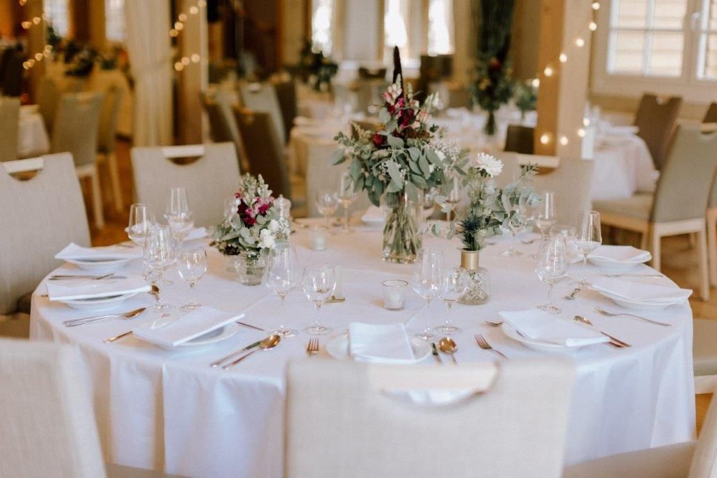 Ein runder Hochzeitstisch mit weißer Tischwäsche, eleganten Blumengestecken, Gläsern und gefalteten Servietten.