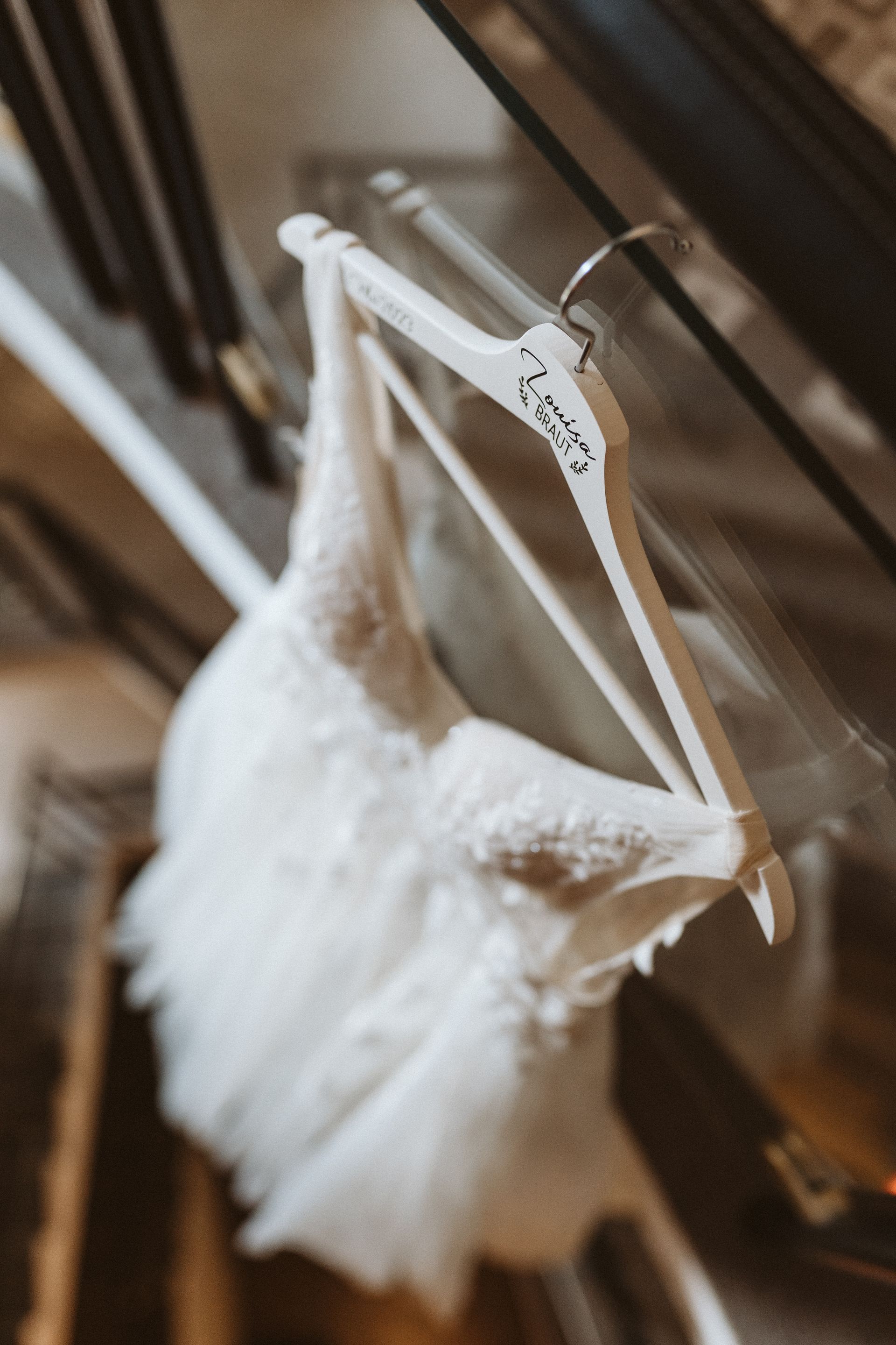 Ein weißes Brautkleid hängt an einem personalisierten Holzkleiderbügel vor einem verschwommenen, dunklen Hintergrund.