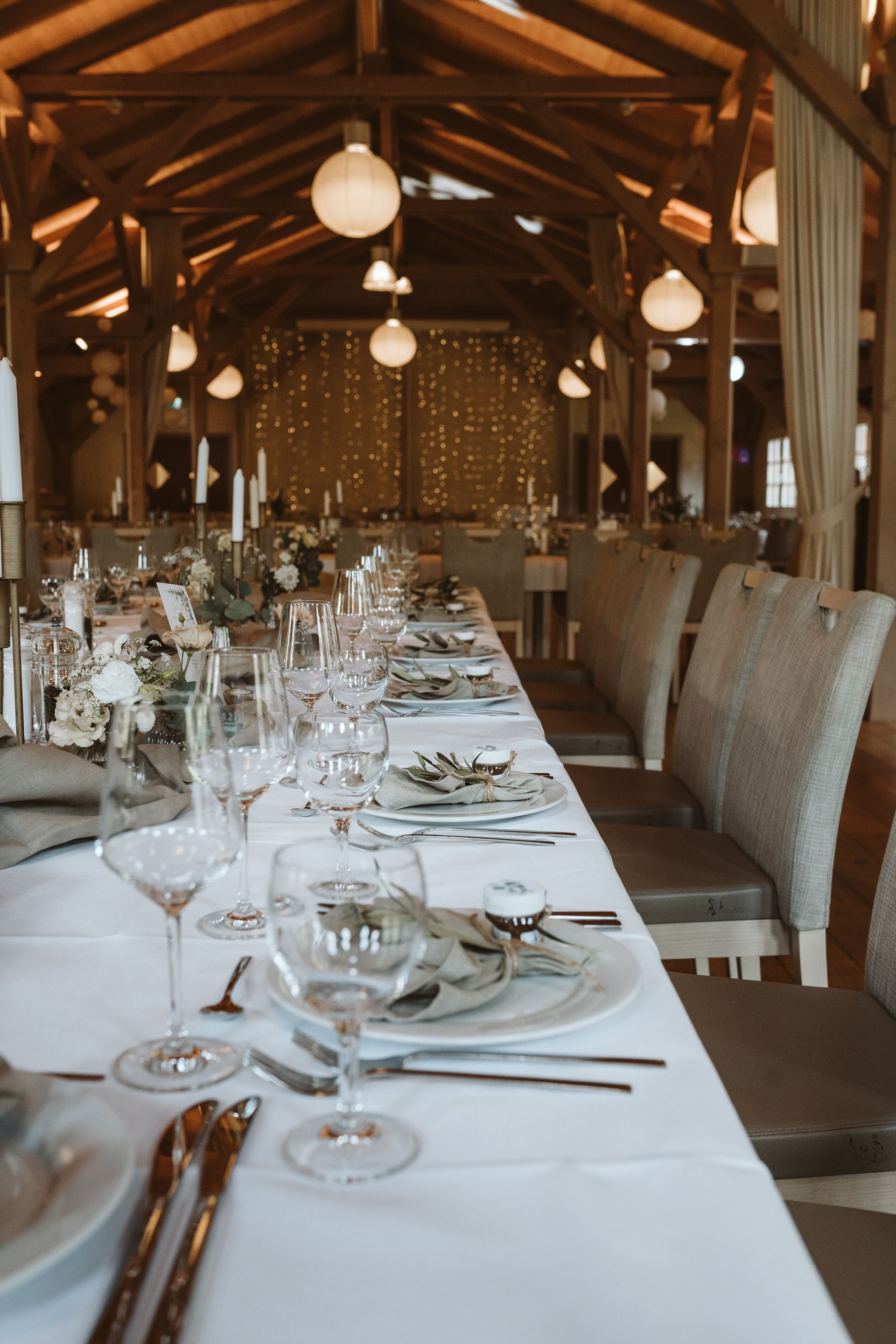 Eine lange Tafel, gedeckt für eine formelle Veranstaltung, in einem rustikalen Ambiente mit Holzbalken und warmen Hängelaternen.