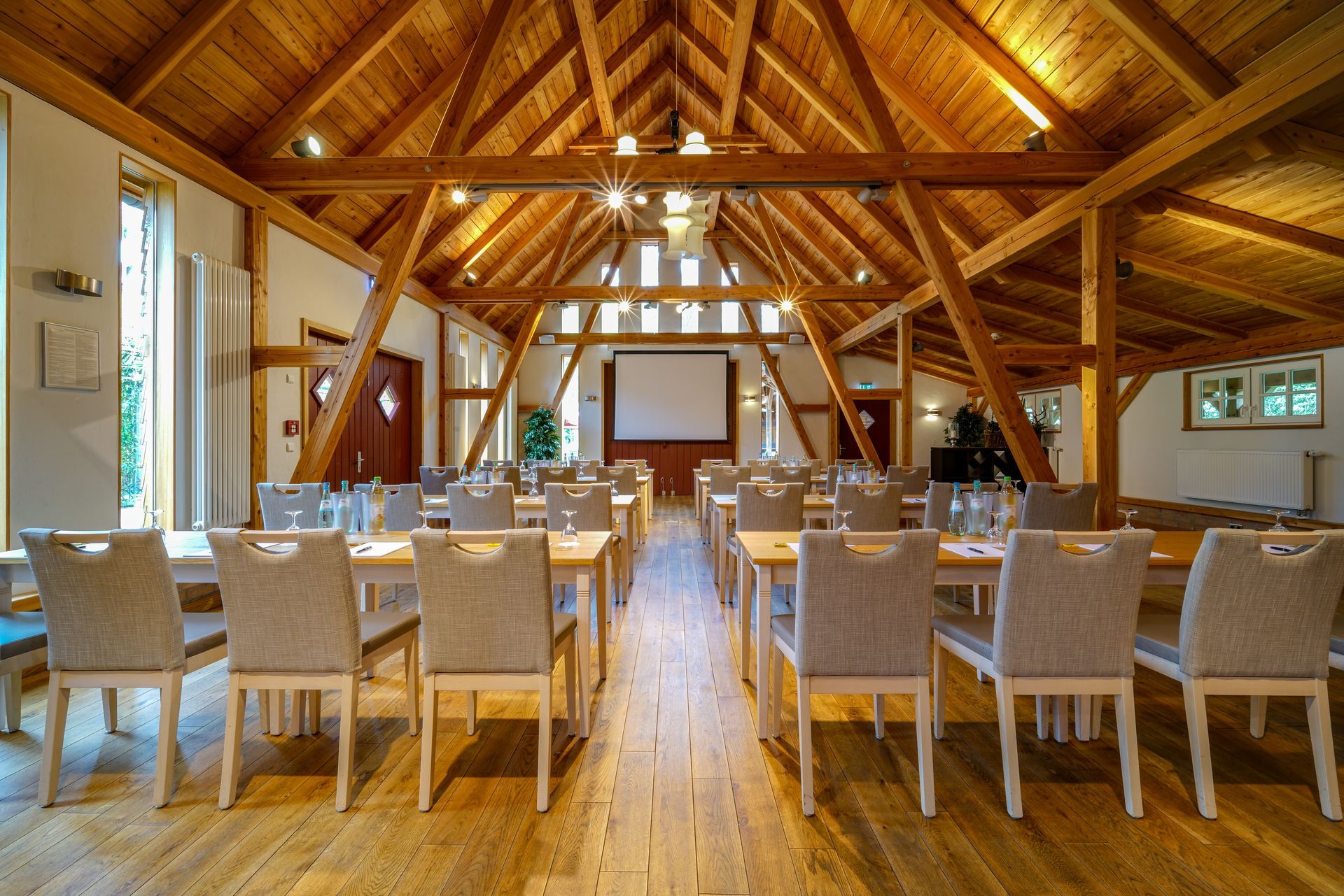 Ein rustikaler Konferenzraum mit hoher Holzbalkendecke, Stuhlreihen, Tischen und einer Präsentationswand an der Vorderseite.