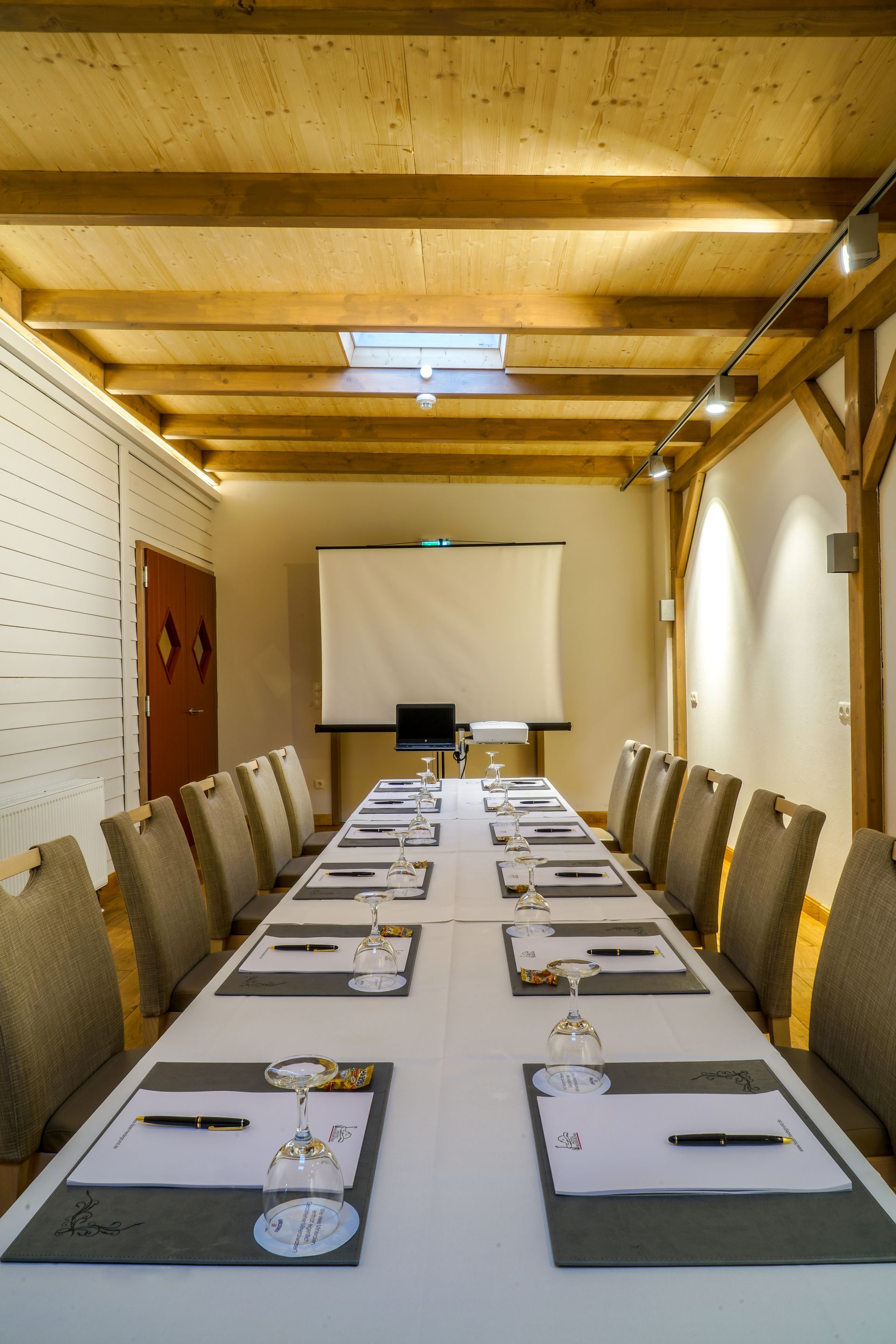 A conference room with a long table, white chairs, and a projection screen beneath a wooden beam ceiling.