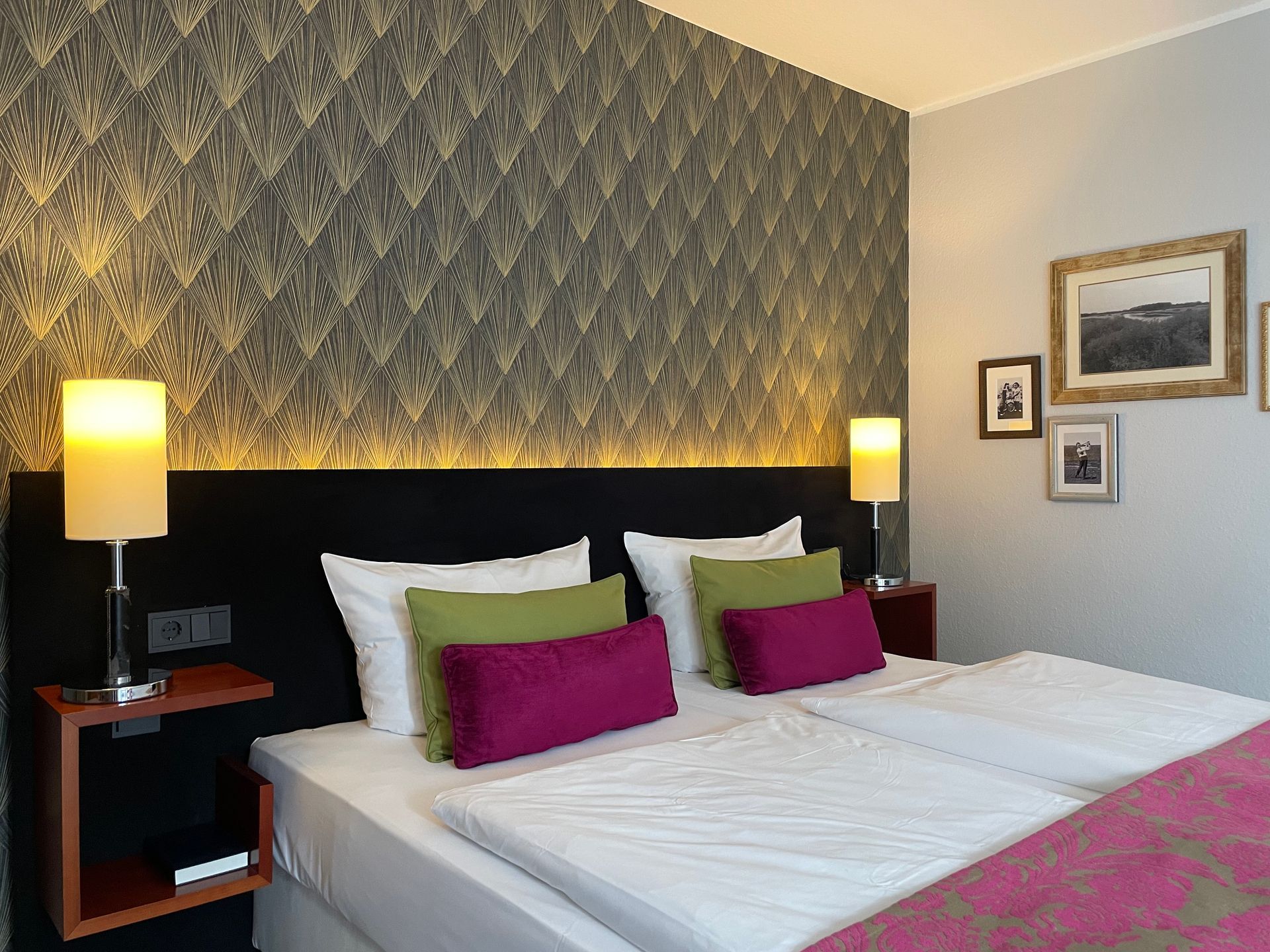 Ein modernes Hotelzimmer mit einem ordentlich gemachten Doppelbett, dunkel gemusterten Kissen, Wandlampen und einer kleinen Sitzecke.