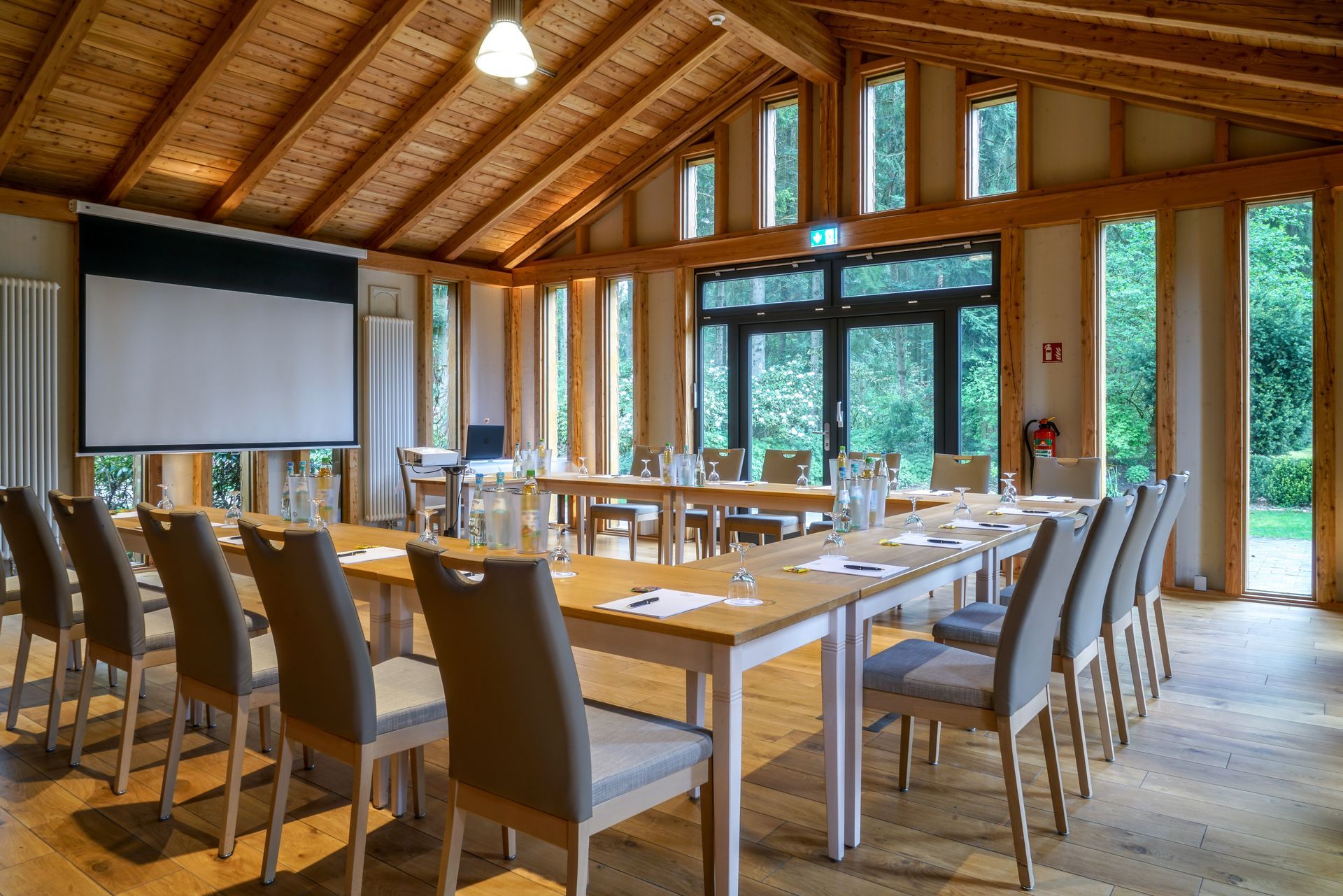 Ein Konferenzraum mit gewölbter Holzdecke, U-förmigen Tischen, grauen Stühlen und einer Projektionsleinwand.
