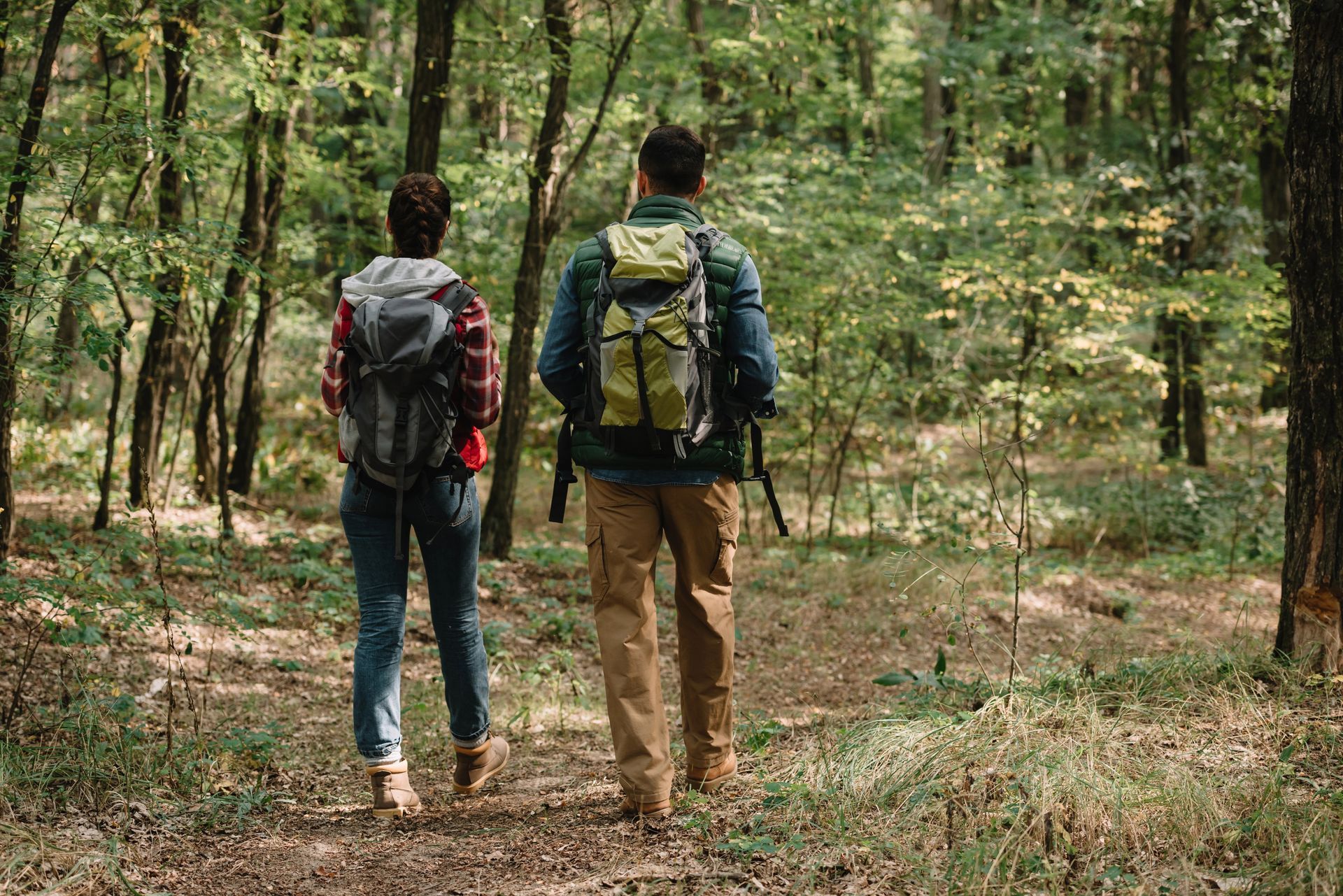Two hikers with backpacks are walking along a trail through a sunlit forest.