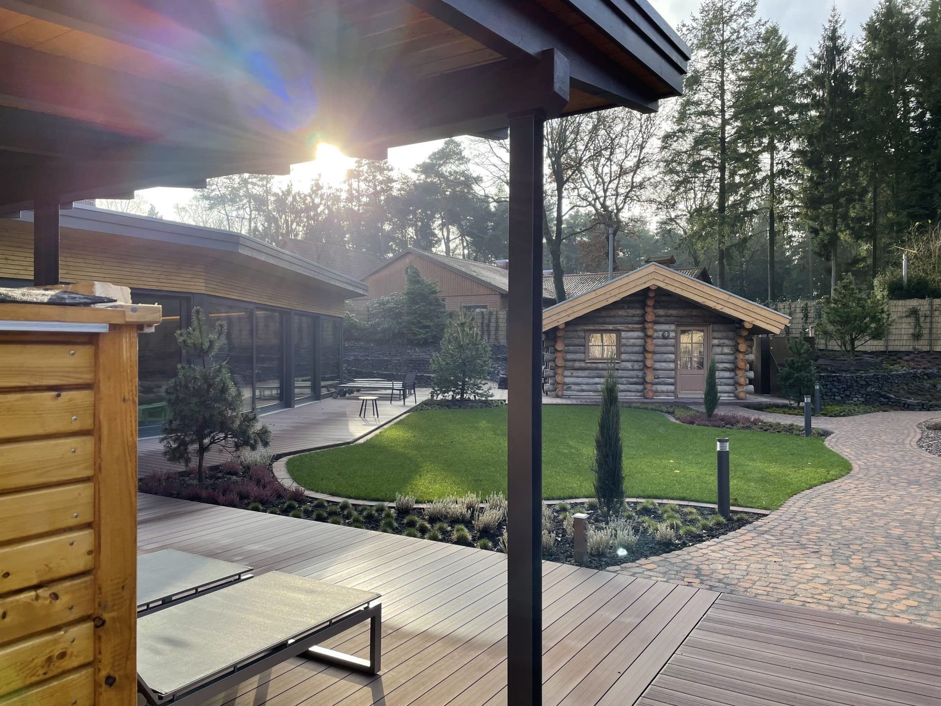 Ein malerischer Garten mit Holzterrasse, gepflegtem Rasen, Steinwegen und einer kleinen Blockhütte unter strahlender Sonne.