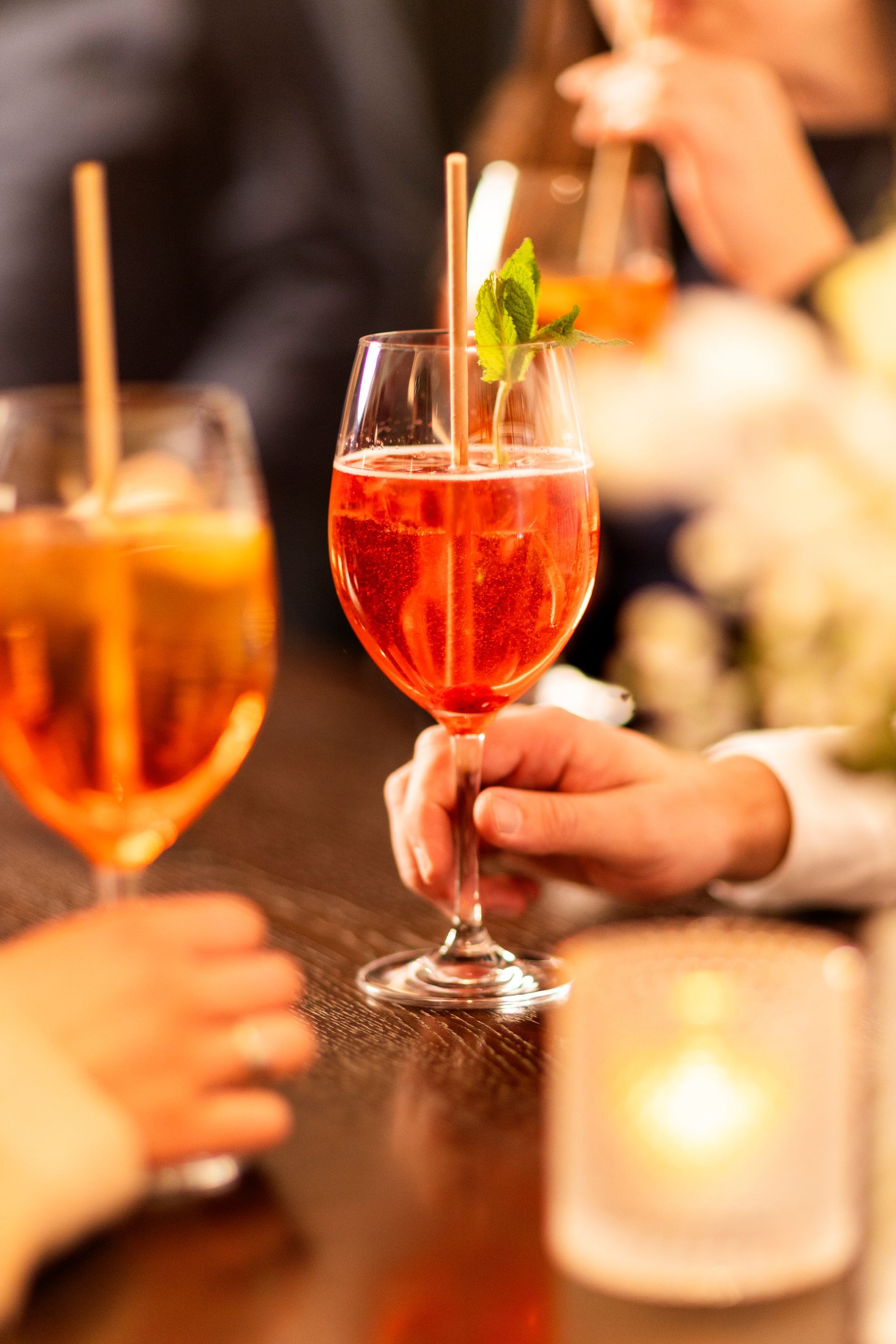 Close-up of hands holding two orange and red spritzer cocktails with straws and mint garnishes on a table with a candle.