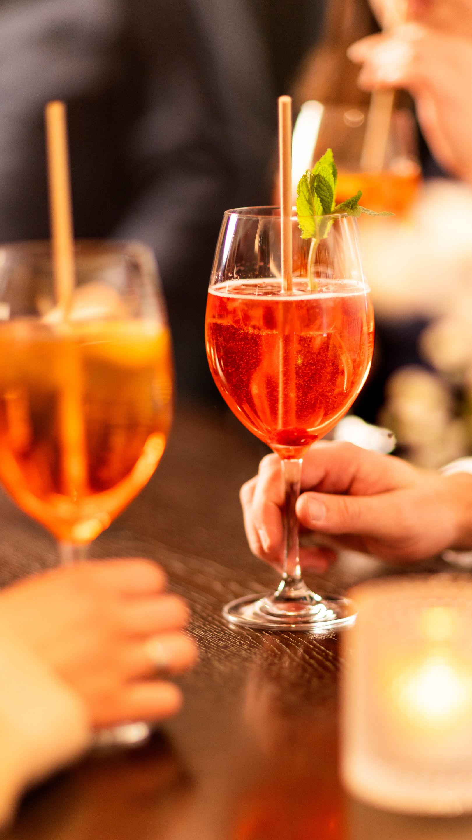 Two hands are holding glasses of orange cocktails, complete with straws and mint garnishes, on a table in a dimly lit setting.