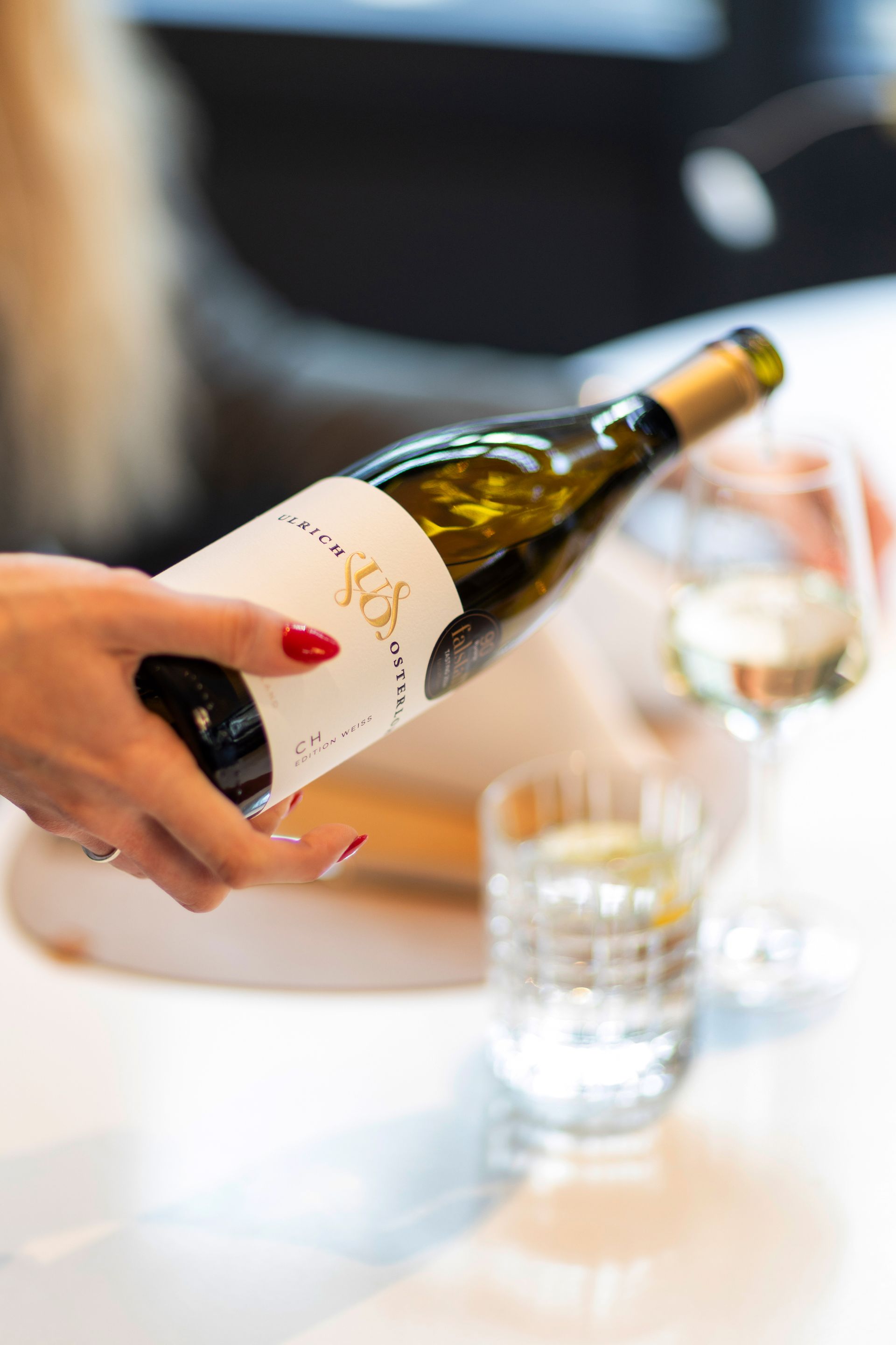 A hand pours white wine from a glass bottle into a stemless wine glass on a table.