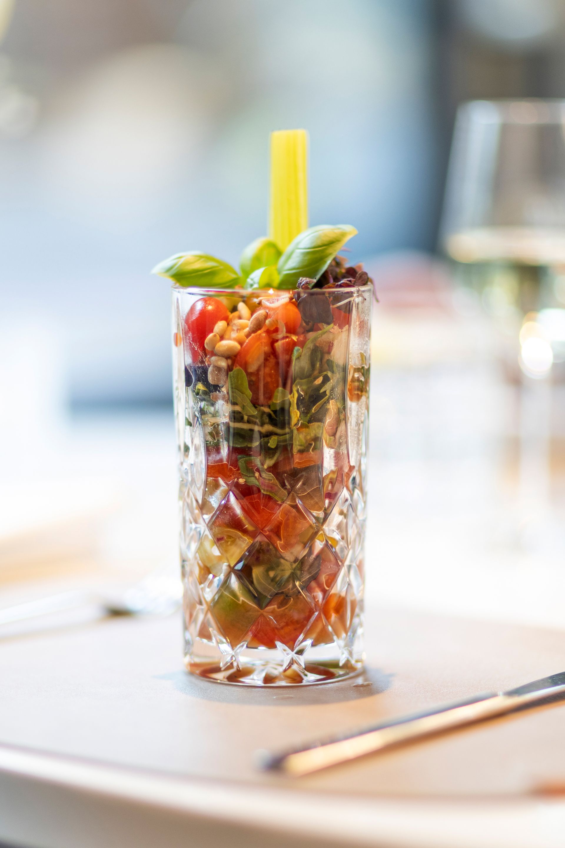 A salad of chopped tomatoes, leafy greens, and pine nuts served in a textured crystal glass, garnished with basil and celery.