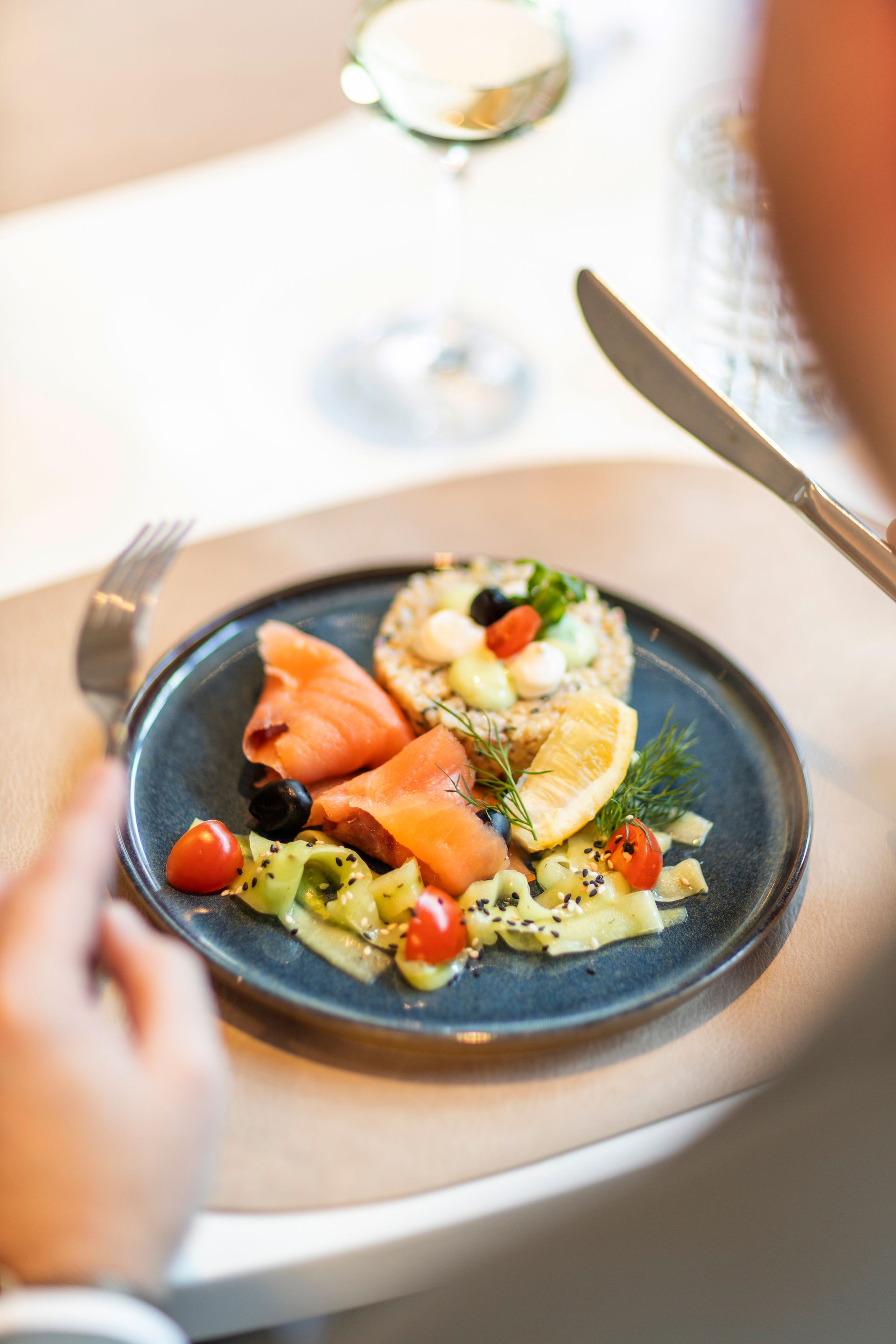 A person is getting ready to eat a gourmet plate of smoked salmon, grains, vegetables, and lemon at a dining table.