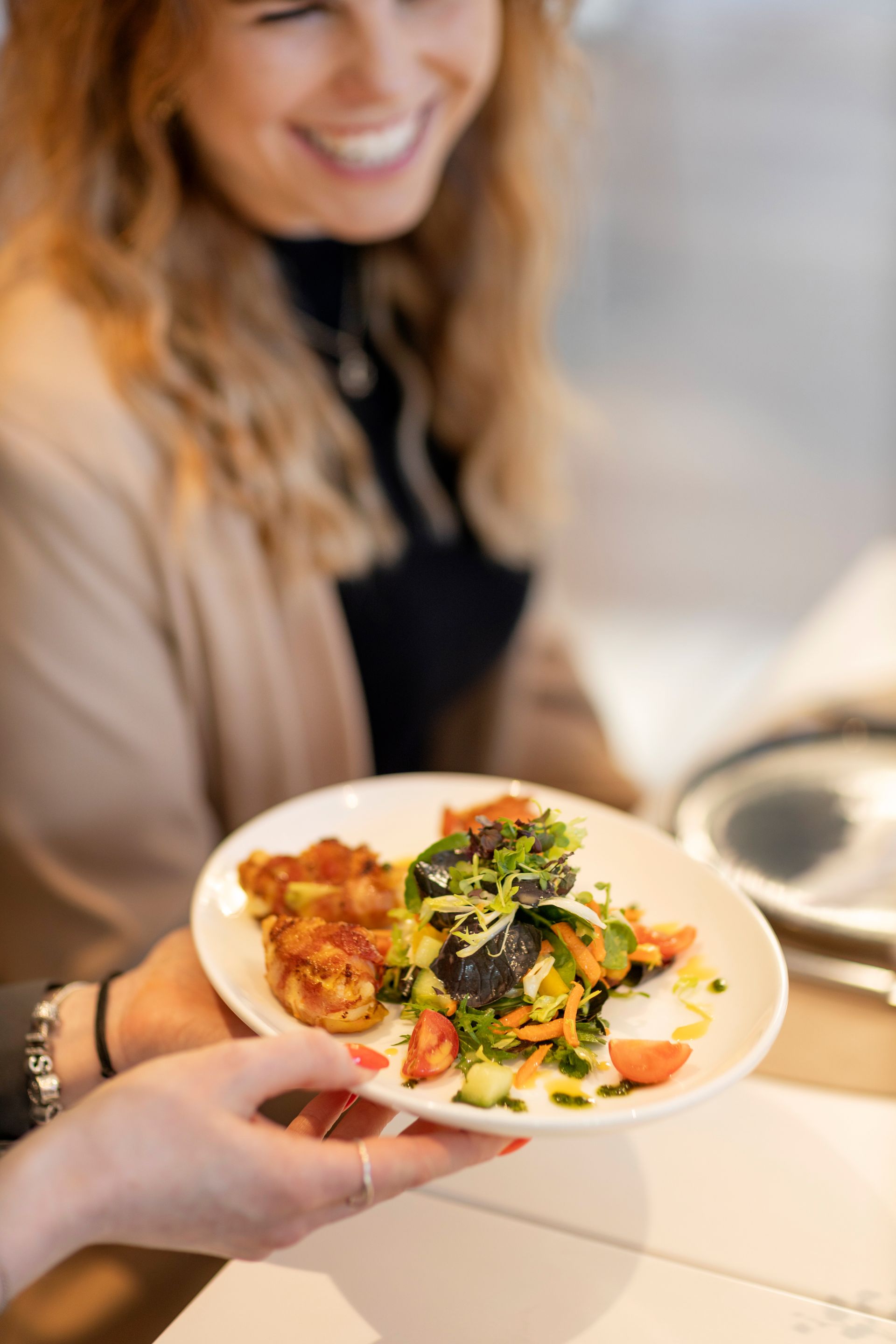 A person is holding a white plate with a fresh salad made of chopped tomatoes and leafy greens; next to them, a smiling person can be seen in the background.