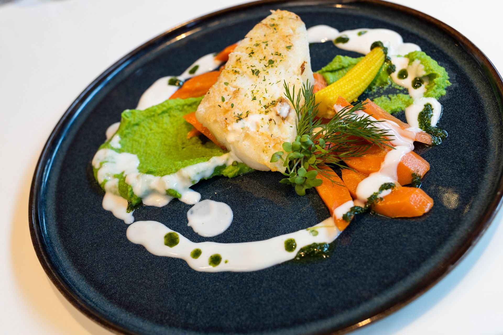 Ein angebratenes Weißfischfilet, serviert mit Erbsenpüree, gerösteten Karotten und einem Spritzer weißer Sauce auf einem dunklen Teller.