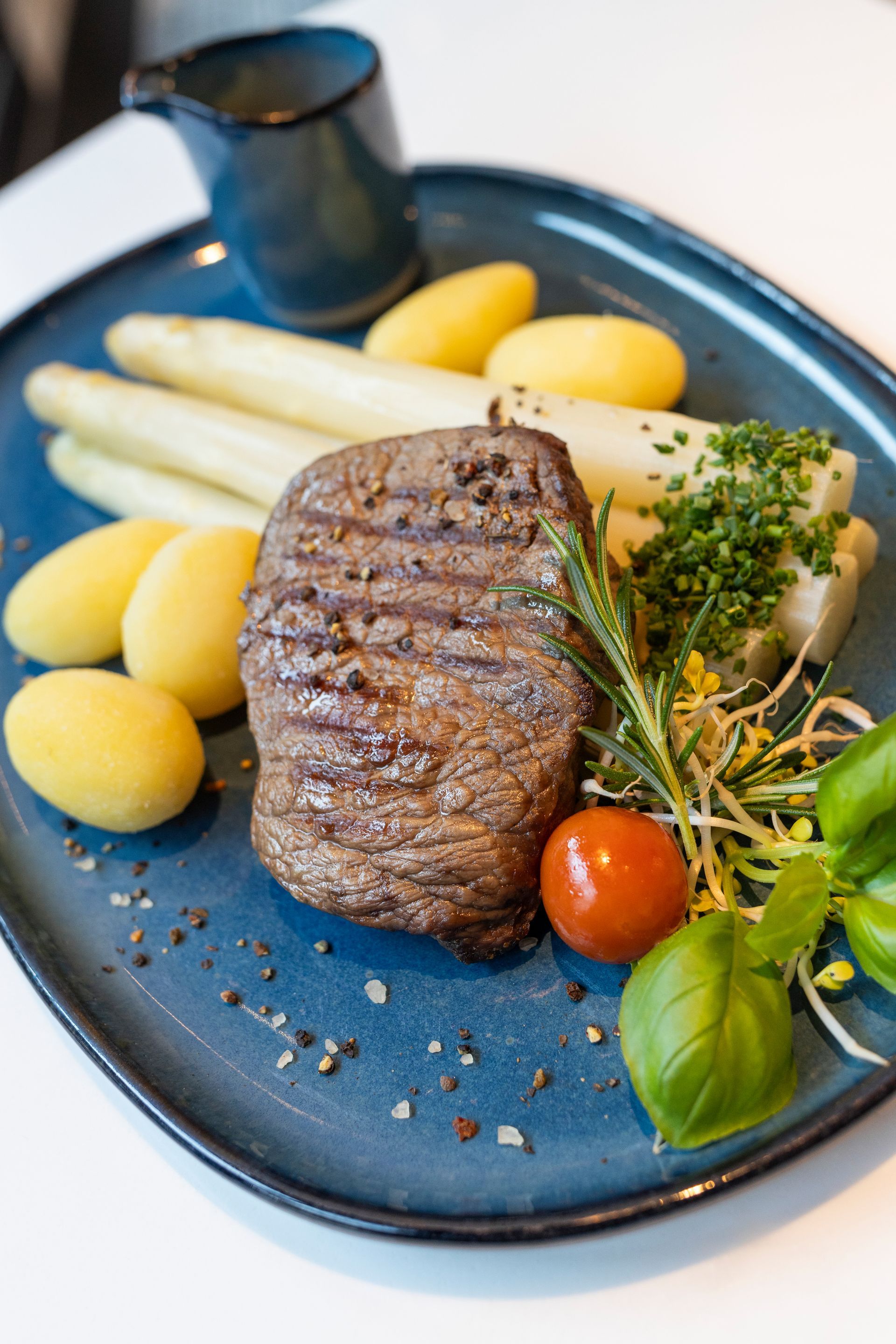 Een gegrilde steak, geserveerd op een blauwe schaal met asperges, gekookte aardappelen, een saus en verse kruiden.