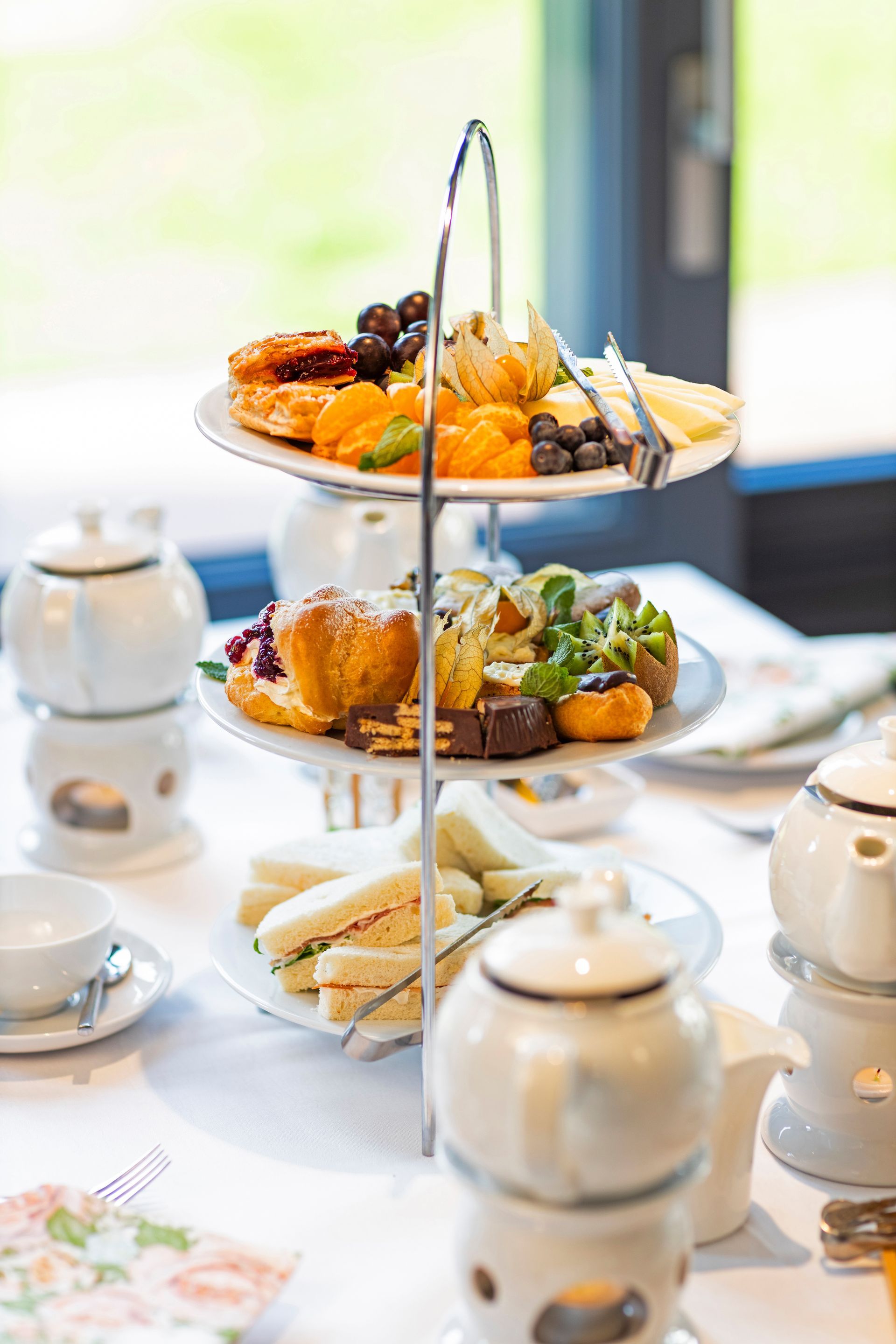 A three-tiered cake stand, filled with various pastries, sandwiches, and fruit, stood on a table alongside teapots.