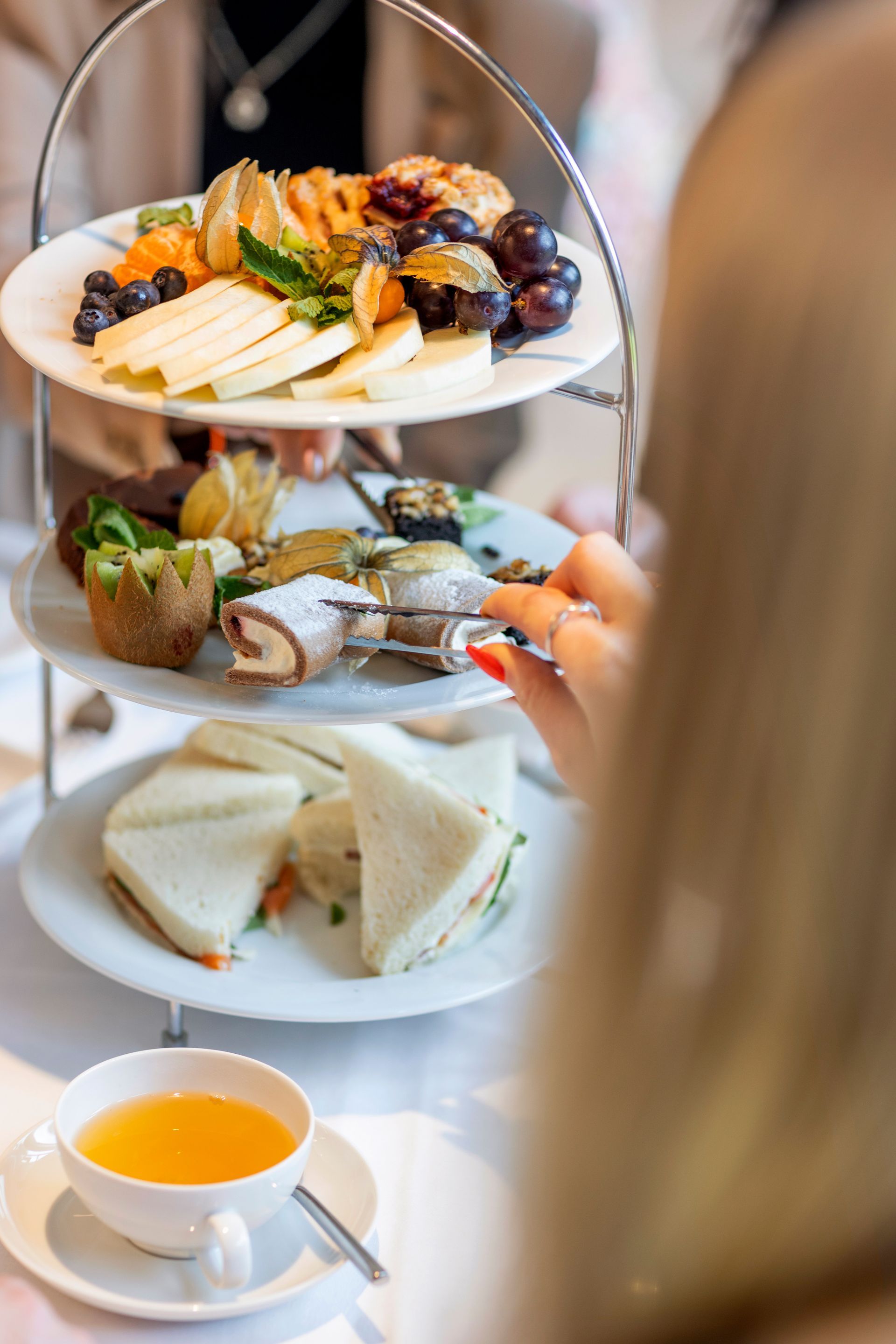 Eine Person greift nach einem Gebäckstück auf einem dreistöckigen Servierständer, der mit Sandwiches, Obst und Süßigkeiten neben Tee gefüllt ist.
