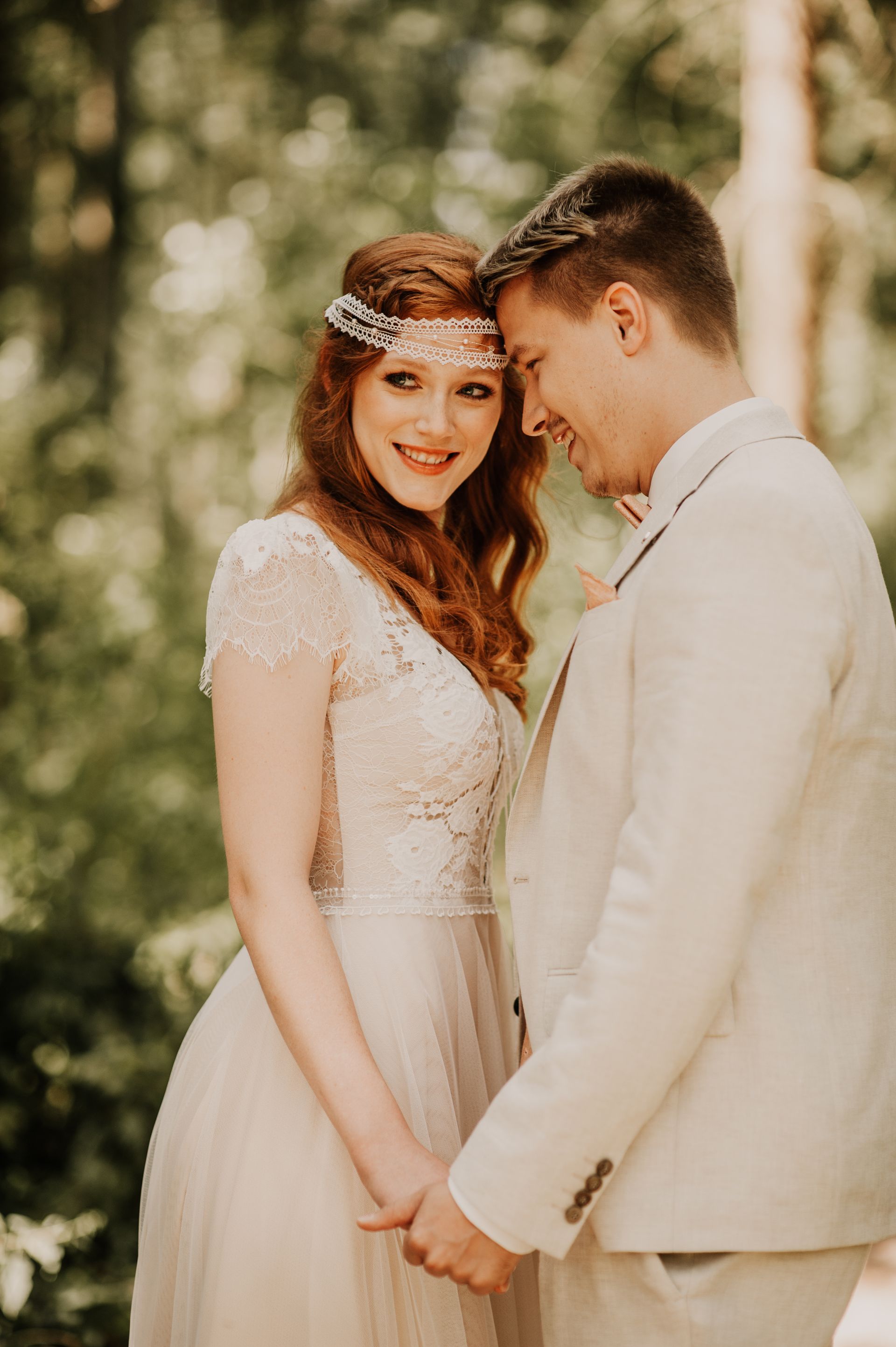 Ein Paar in Hochzeitskleidung hält Händchen und lächelt sich in einer bewaldeten Umgebung an.