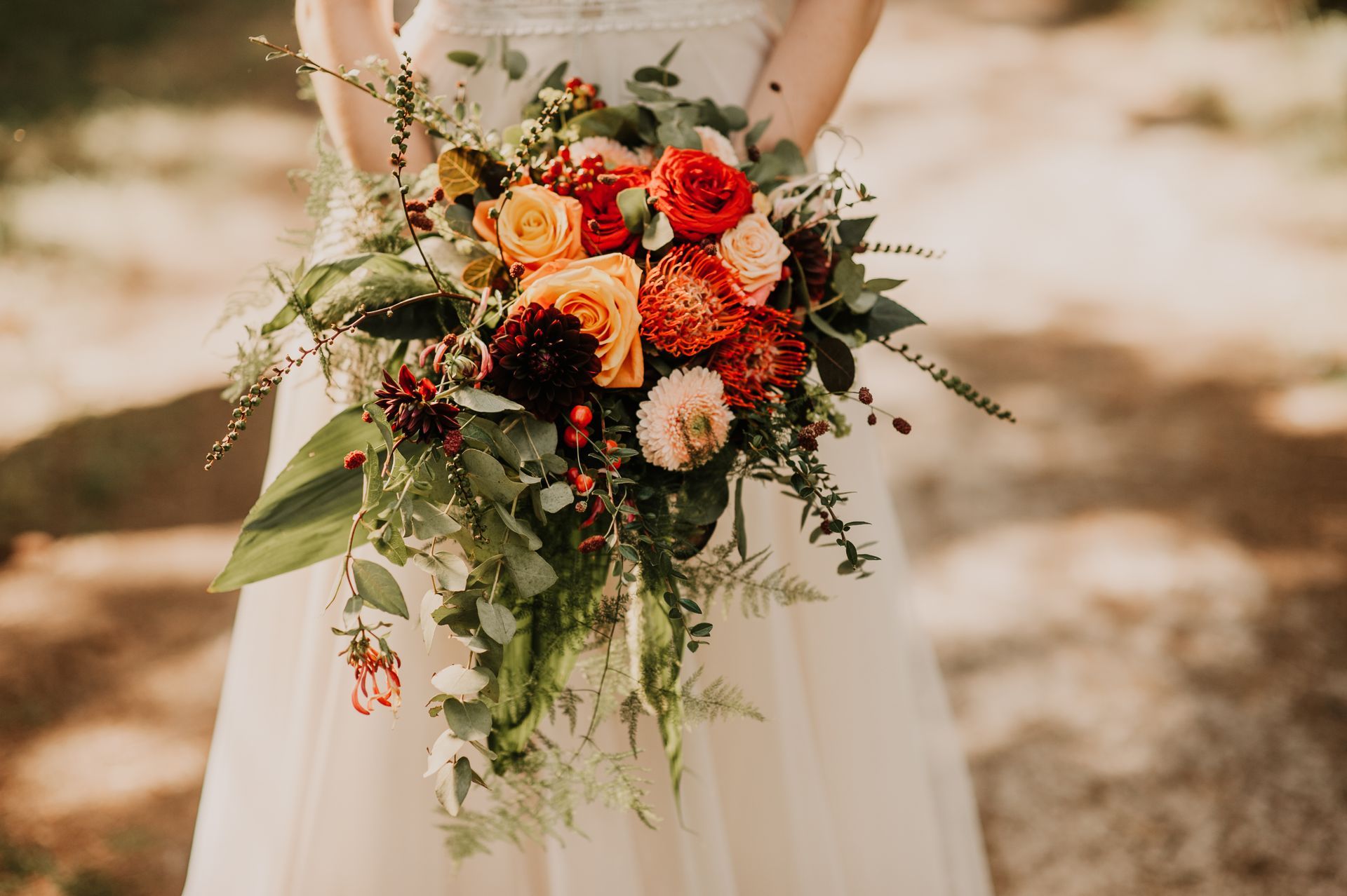 Een bruid houdt in een openluchtdecor een trapsgewijs opgebouwd bruidsboeket vast, bestaande uit oranje, rode en perzikkleurige bloemen met weelderig groen.