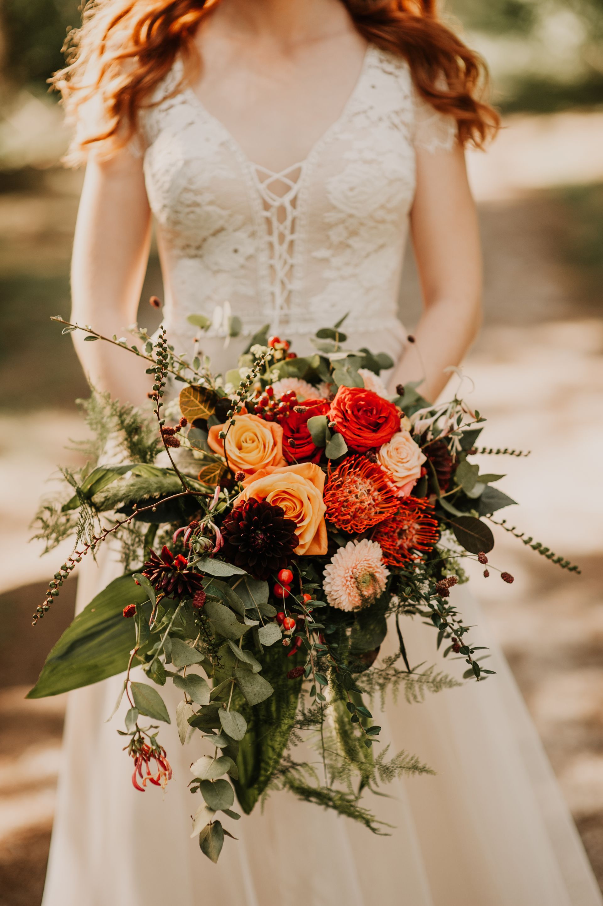 Een vrouw in een witte kanten bruidsjurk houdt een levendig, rustiek bruidsboeket vast met oranje, rode en donkerbordeauxrode bloemen.