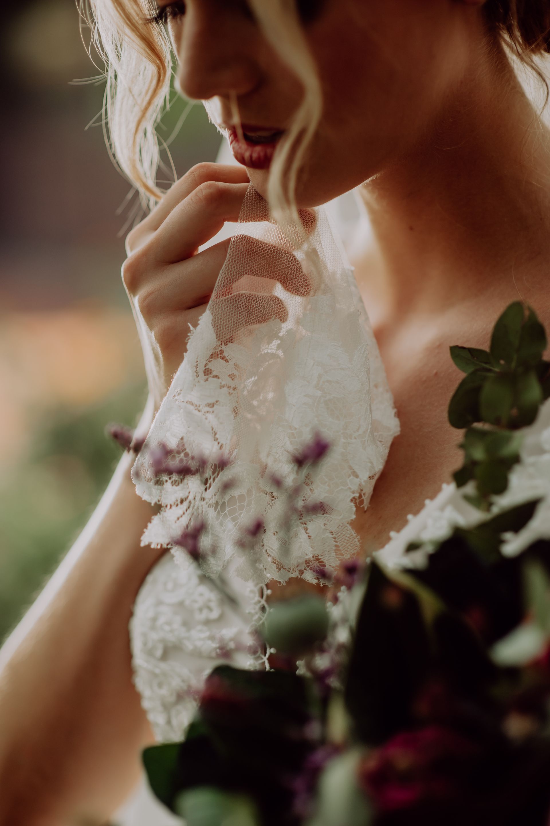 Nahaufnahme einer Person in einem Brautkleid aus Spitze, die einen Brautstrauß hält und sich mit den Fingern sanft ans Kinn fasst.