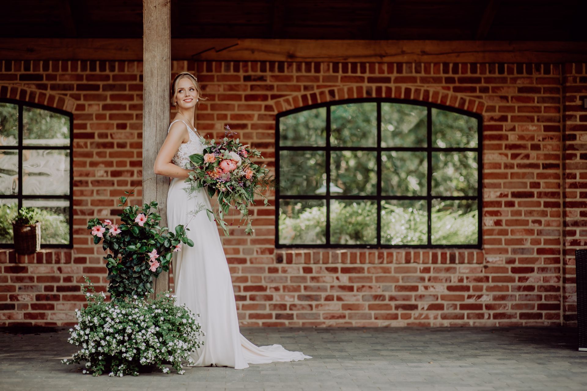 Eine Person in einem weißen Brautkleid hält einen Brautstrauß, während sie in einem Pavillon unter freiem Himmel an einer Backsteinsäule lehnt.