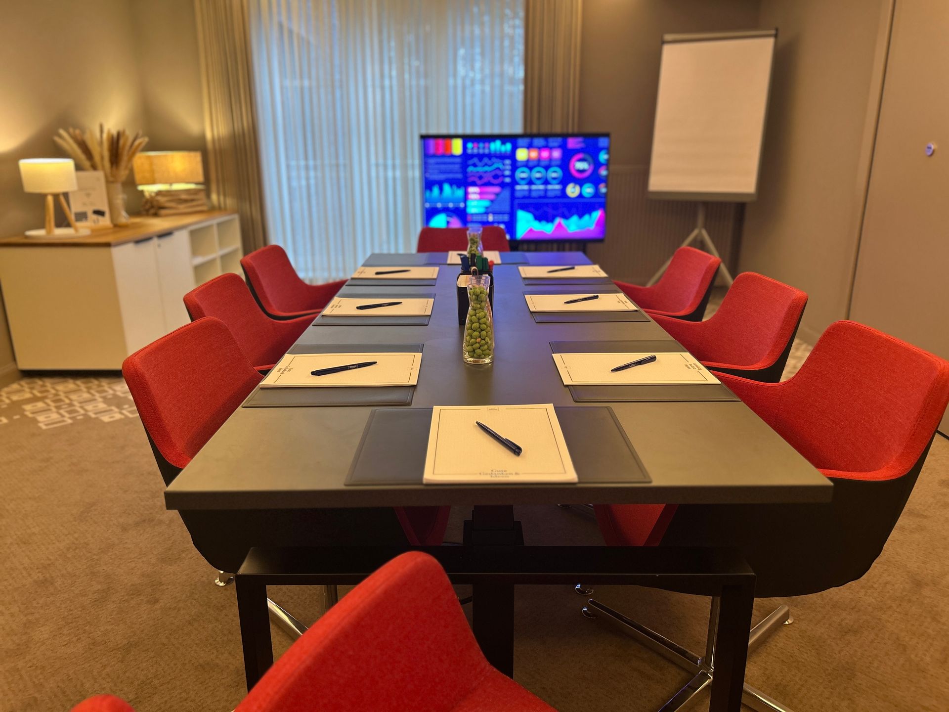 A conference room with a long table, eight red chairs, a monitor displaying data charts, and a whiteboard mounted on a stand.