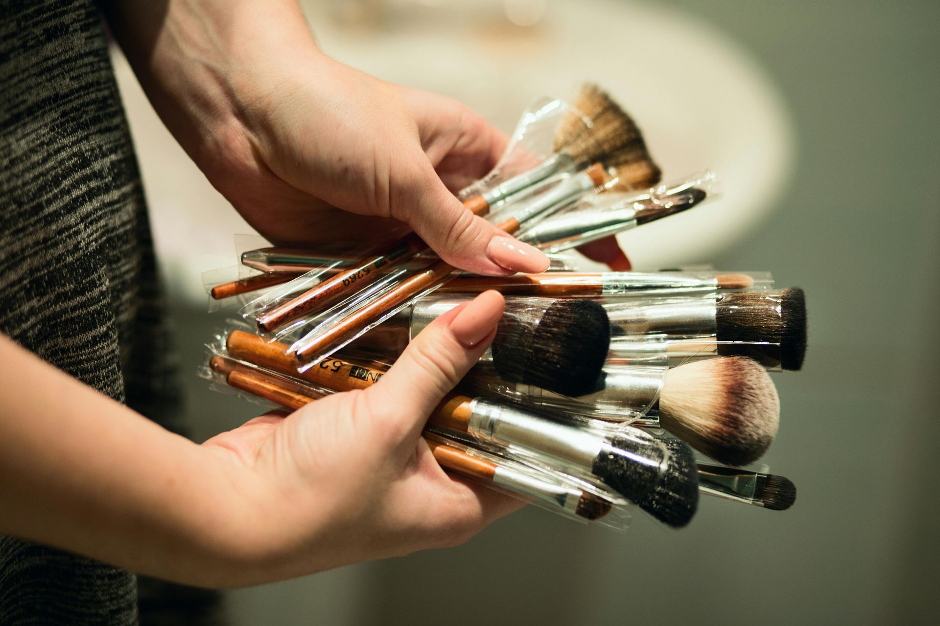 Hands are holding a collection of various makeup brushes with brightly colored handles in a bathroom.