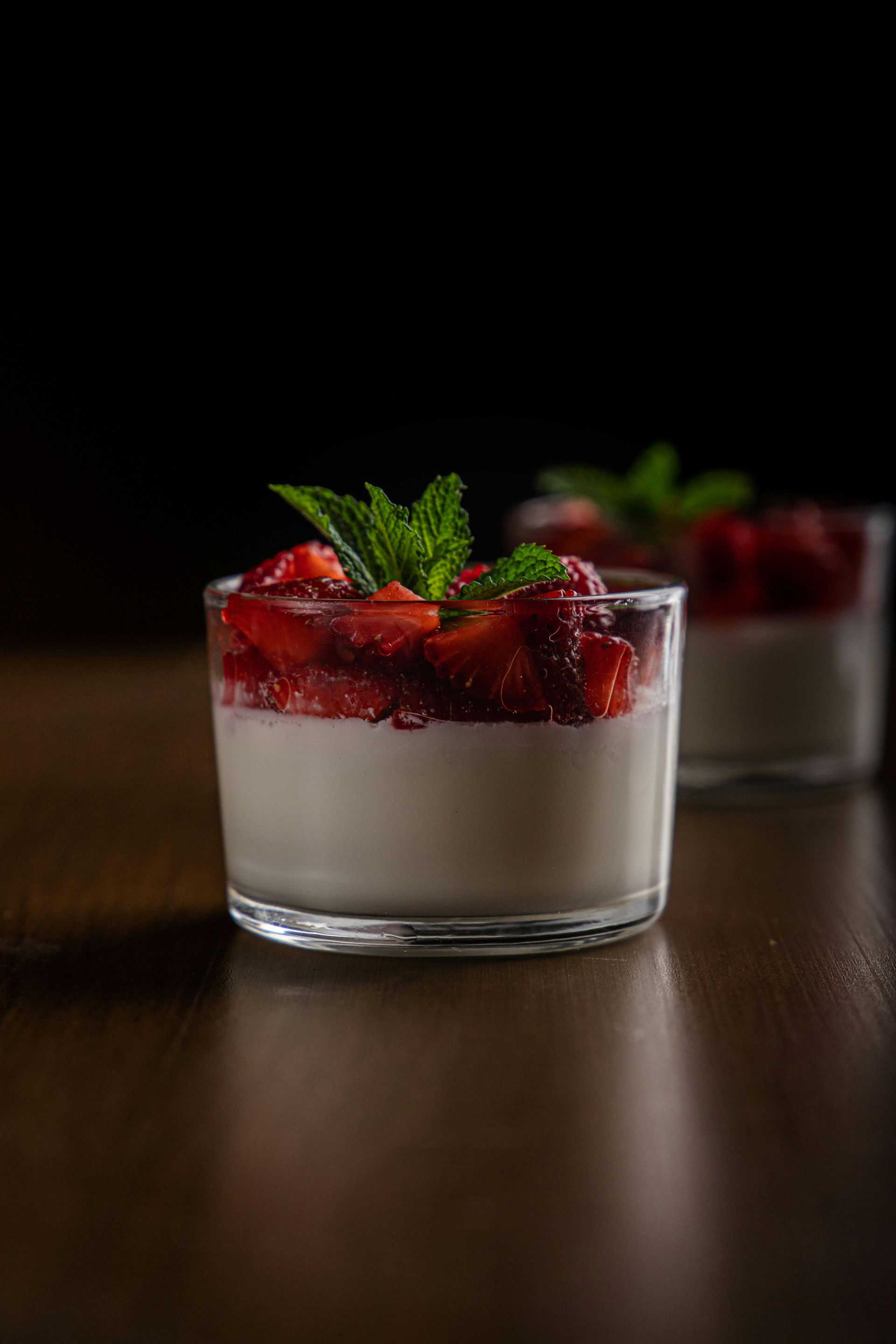 Zwei Gläser Erdbeer-Panna-Cotta, garniert mit frischen Minzblättern, auf einem Holztisch vor dunklem Hintergrund.