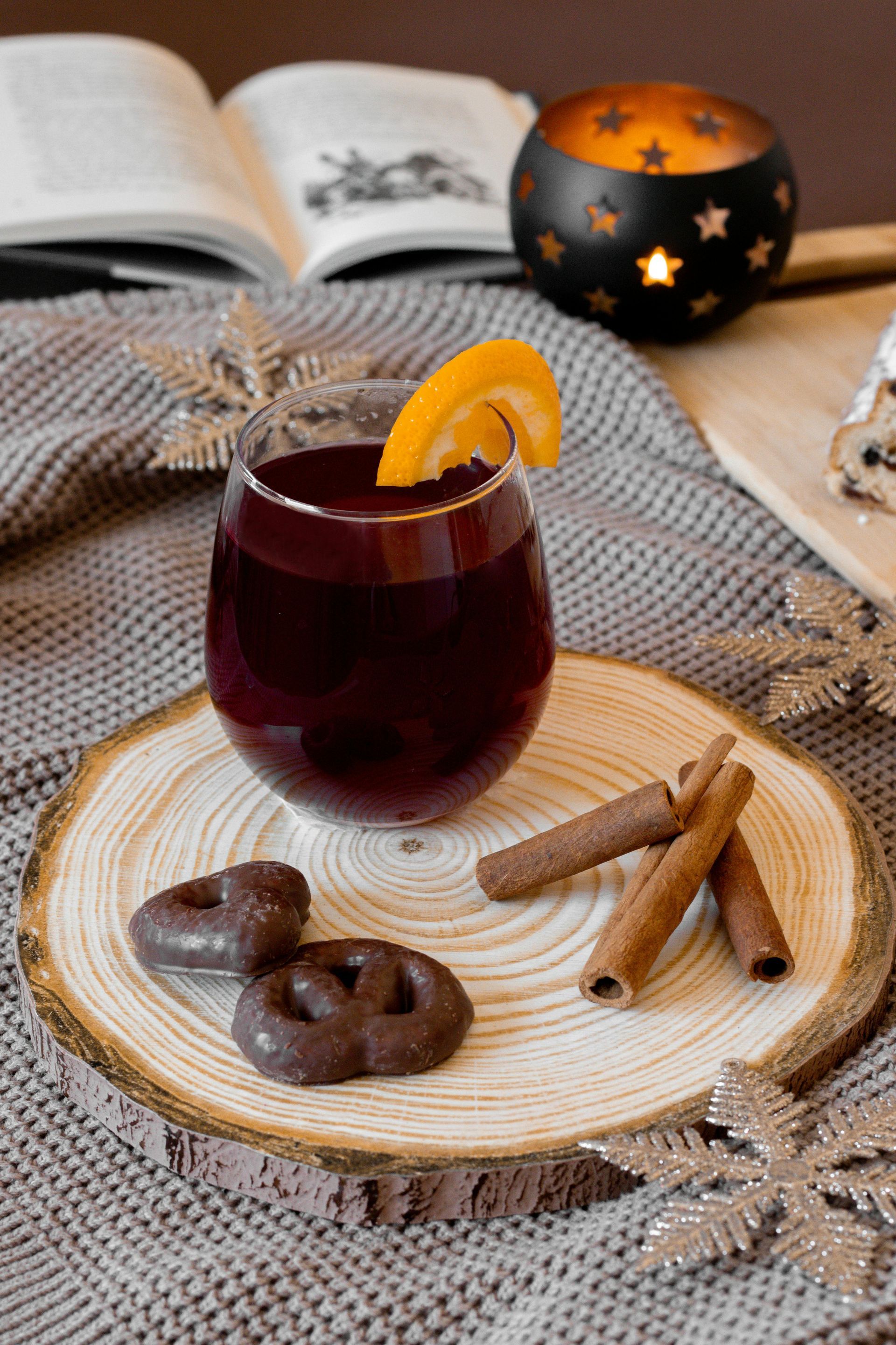 Ein Glas roter Glühwein mit einer Orangenscheibe, Schokoladenbrezeln und Zimtstangen auf einem Holzbrett.