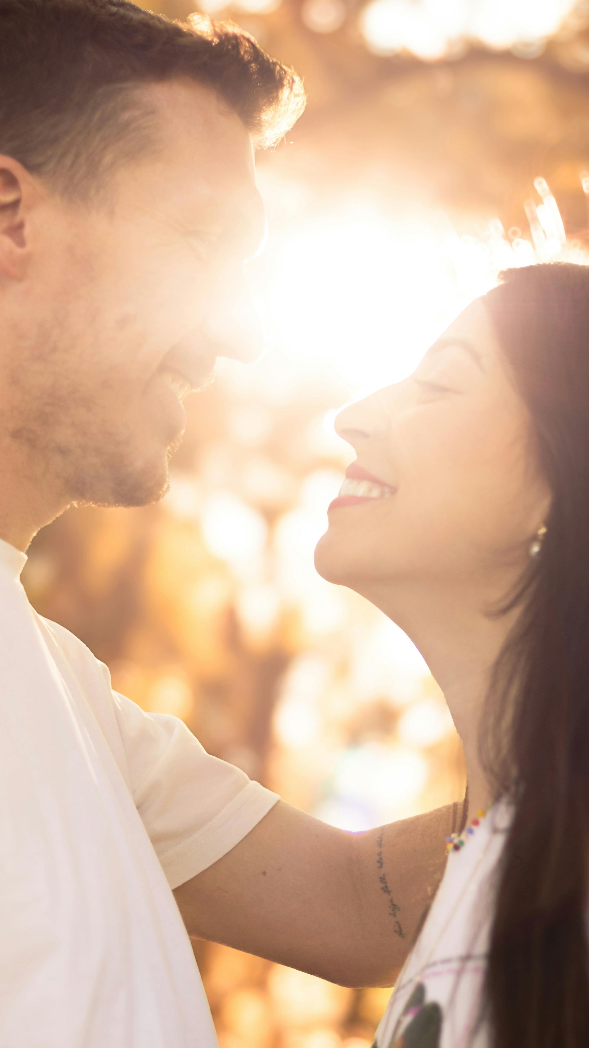 A couple gazes at each other with gentle smiles, bathed in the warm, golden light of the sunset against an outdoor backdrop.
