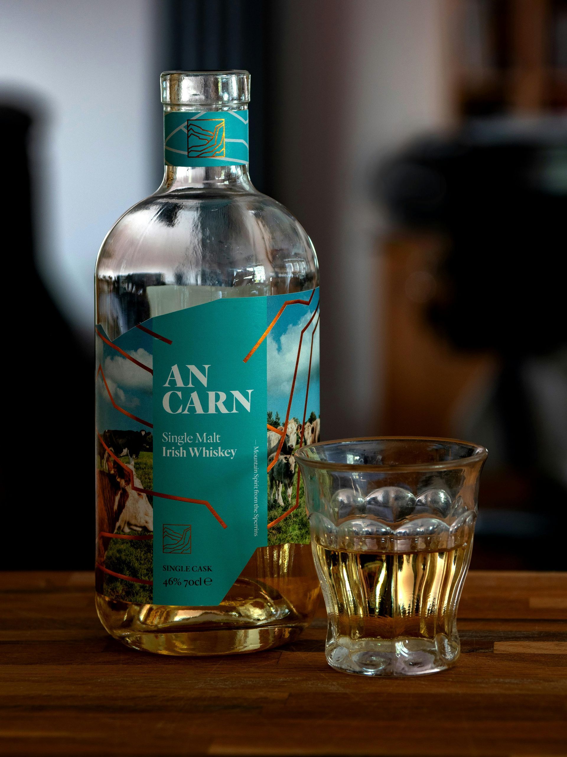 Een fles An Carn Irish Whiskey en een klein glaasje met amberkleurige schnaps op een houten tafel.