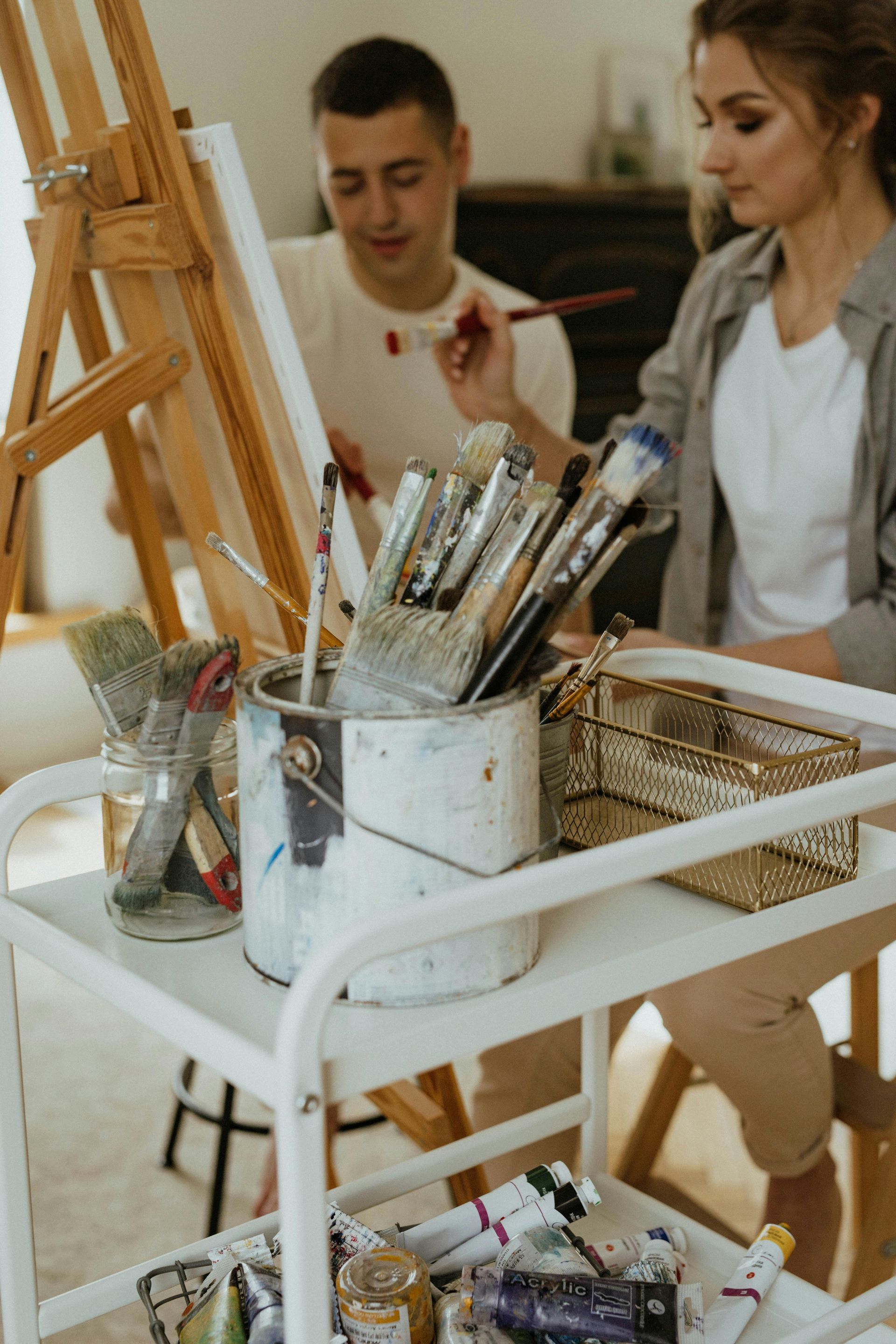 Twee kunstenaars schilderen aan schildersezels in een atelier; op de voorgrond staat een wit karretje met penselen en verfbuisjes.