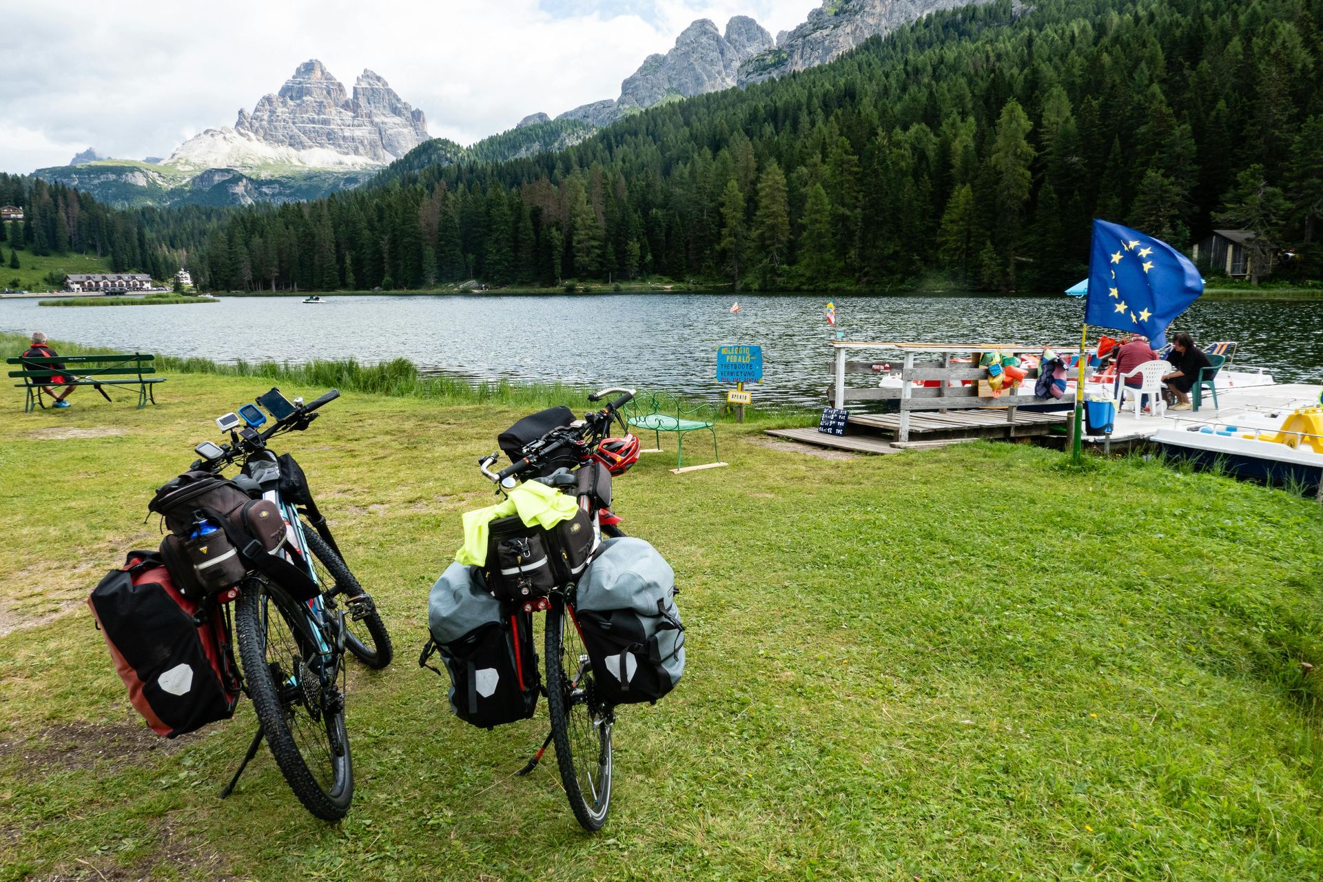 Zwei beladene Tourenräder stehen auf einer Wiese vor einem See; im Hintergrund erheben sich Berggipfel und die Flagge der Europäischen Union.