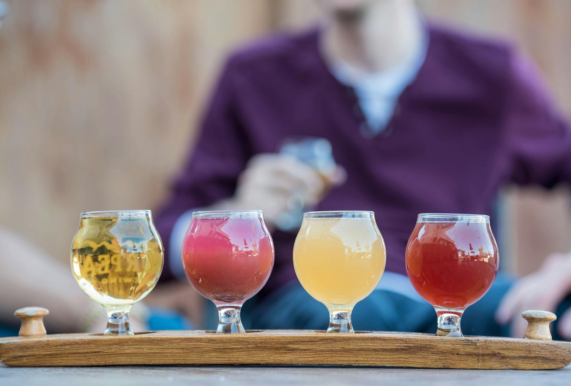 Een selectie van vier kleurrijke craftbieren in bolvormige glazen, gerangschikt op een houten plank, met daarnaast een wazige persoon.
