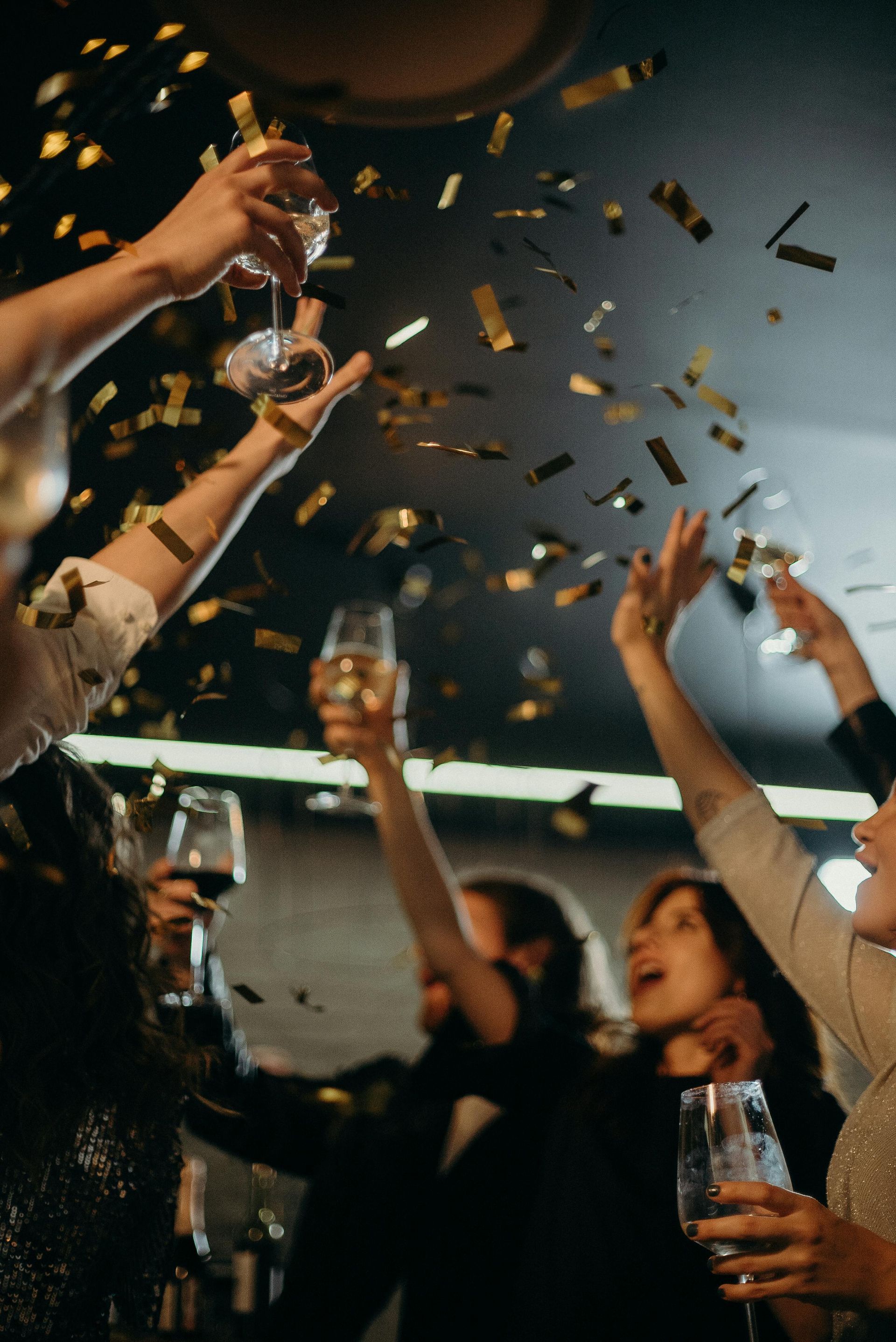 A group of people are cheering with raised glasses amid a shower of gold confetti, creating a joyful, festive atmosphere.