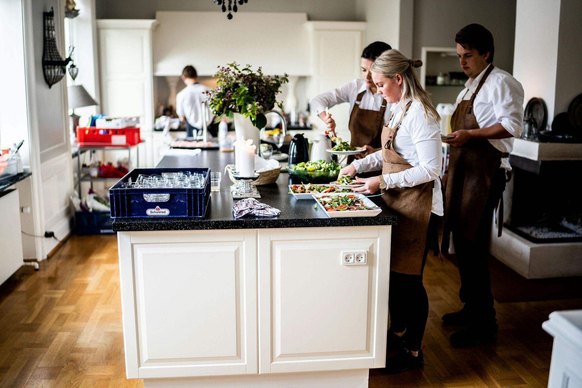 Drie mensen in witte overhemden en bruine schorten staan in een licht, modern huis op een kookeiland borden met eten klaar te maken.