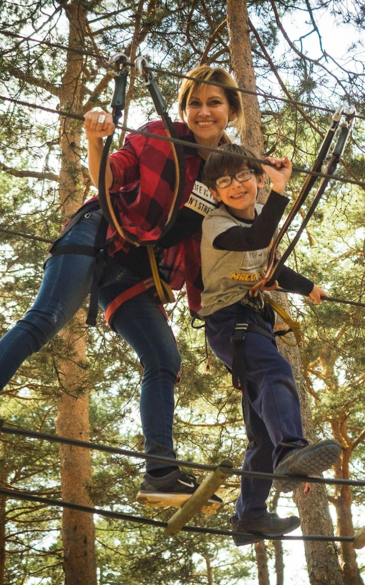 A smiling adult and a child are navigating a high ropes course in a forest, secured by ropes and wearing safety harnesses and equipment.