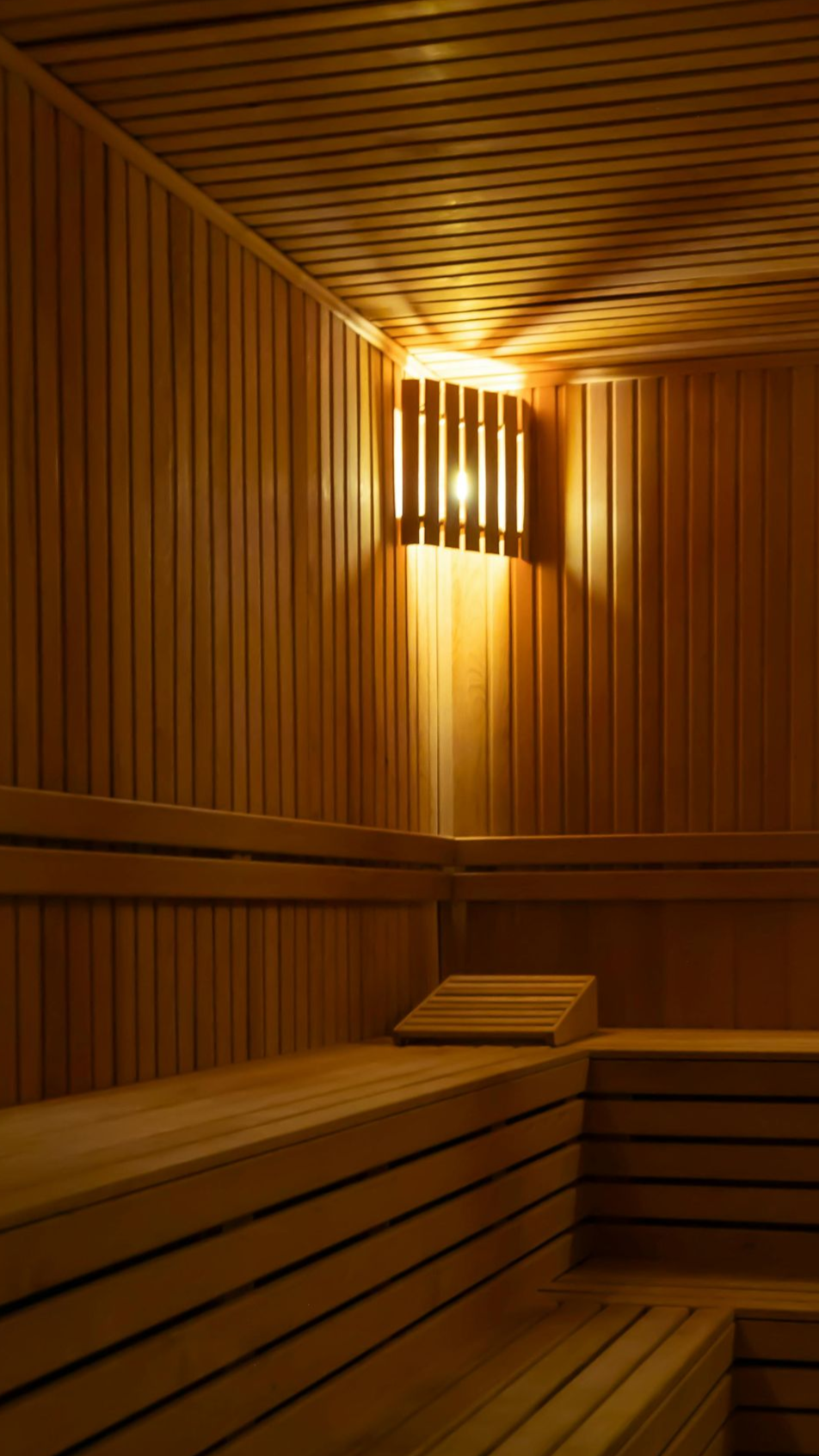 Het interieur van een houten sauna met lattenwanden, getrapte banken en een lamp die aan de hoekwand hangt.