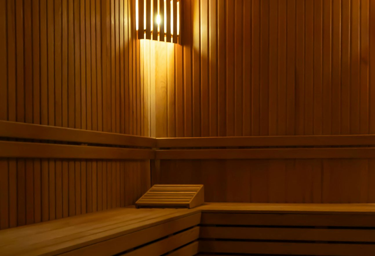 Een hoek van een houten saunacabine met verticale lambrisering, een getrapte bank en een warm schijnende wandlamp.