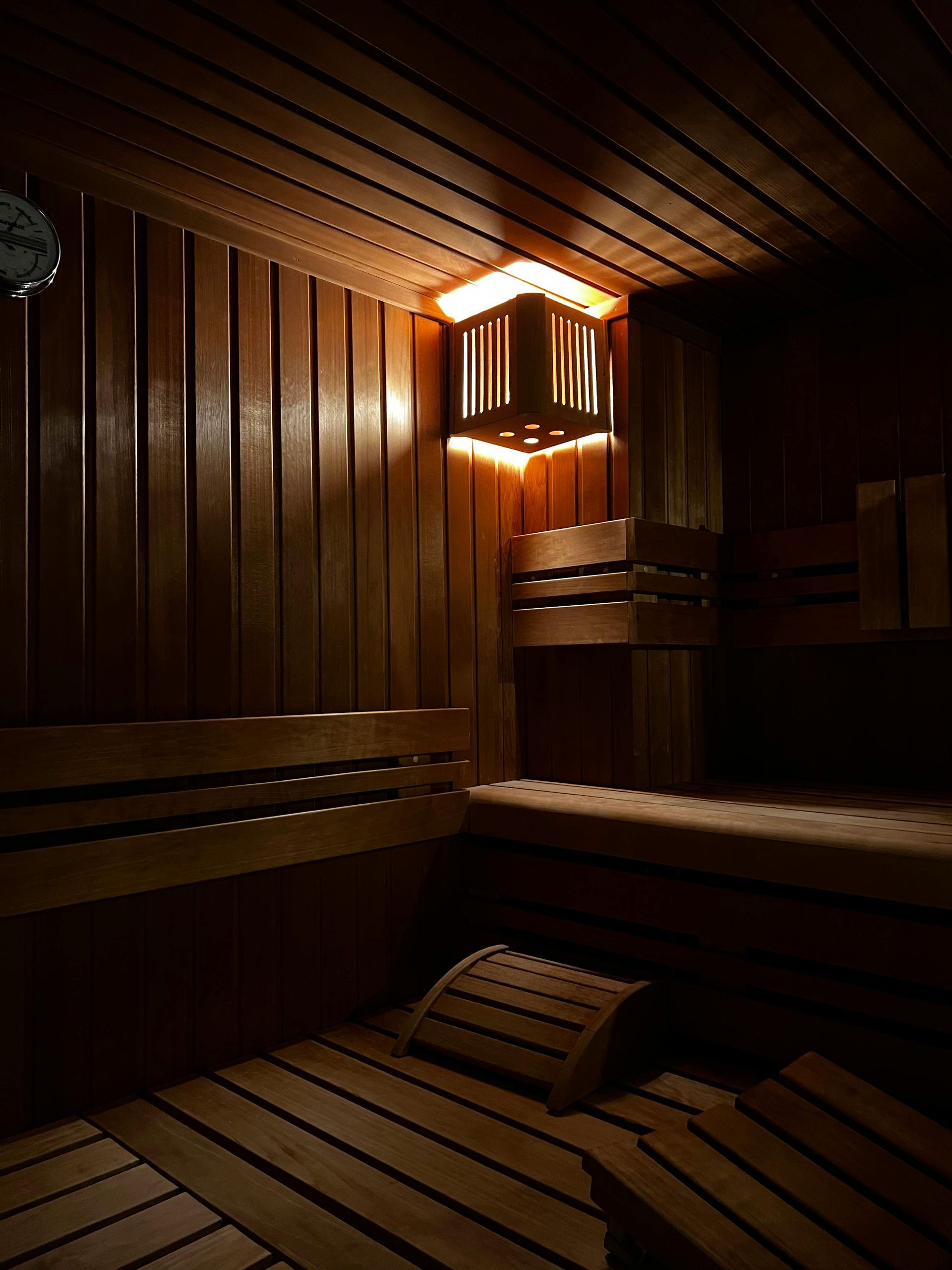 Een schemerig verlichte sauna met warme houten wanden, lattenbanken en een kleine, vierkante wandlamp.