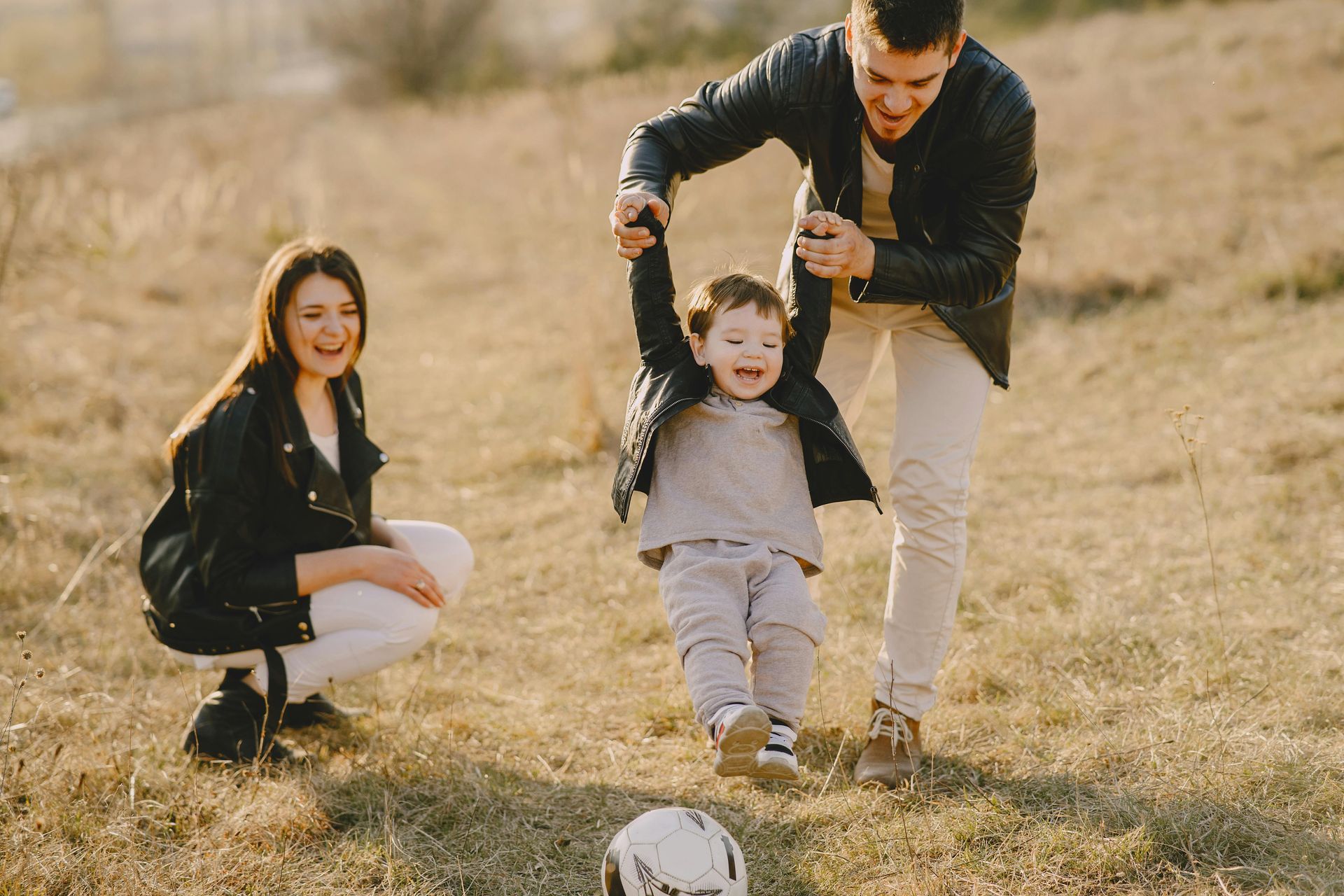 A family is playing with a soccer ball on a dry, grassy field; one parent is holding a child's hands while the other parent watches.