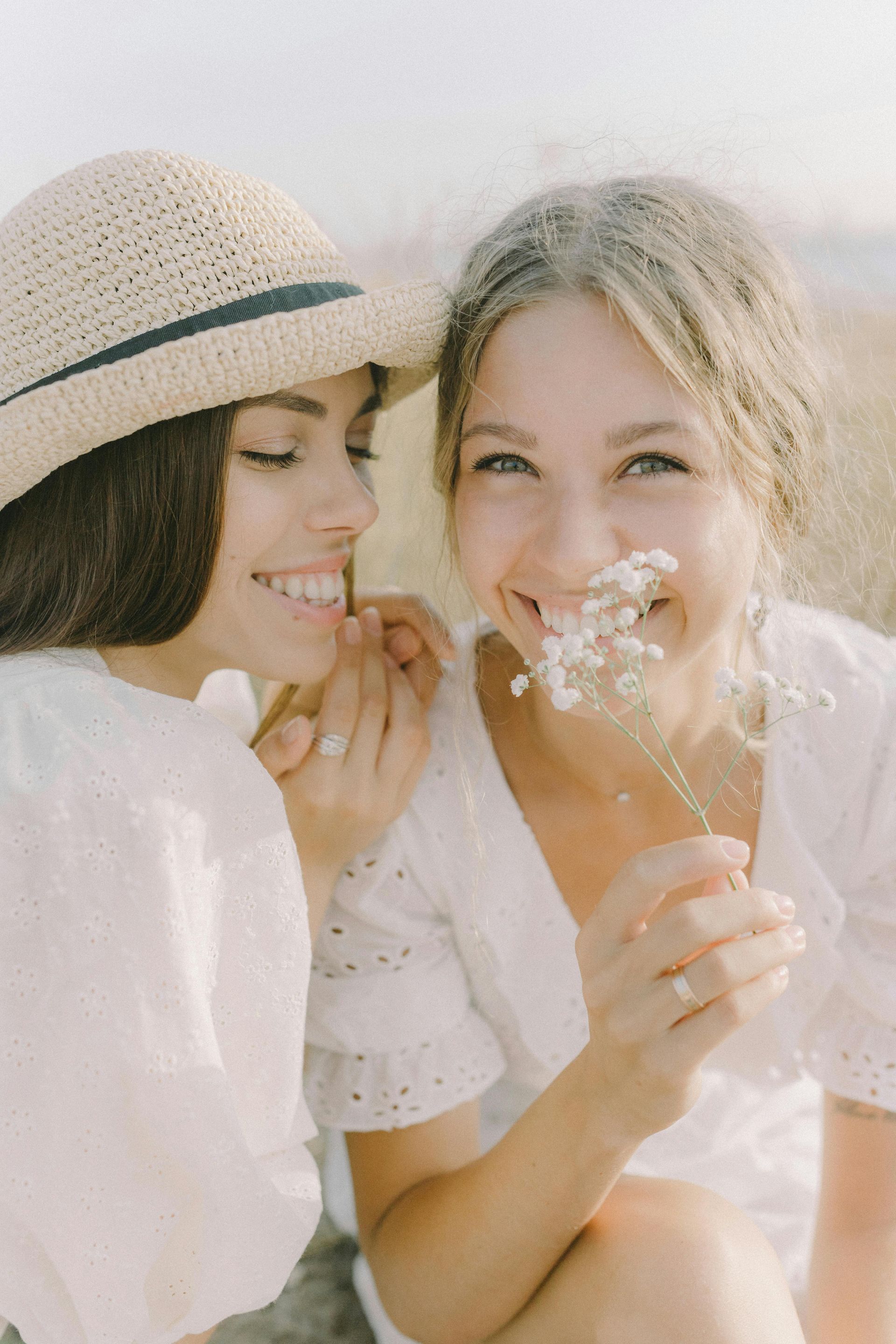 Two smiling people wearing white tops are posing outdoors; one is wearing a straw hat and holding a sprig of white flowers.