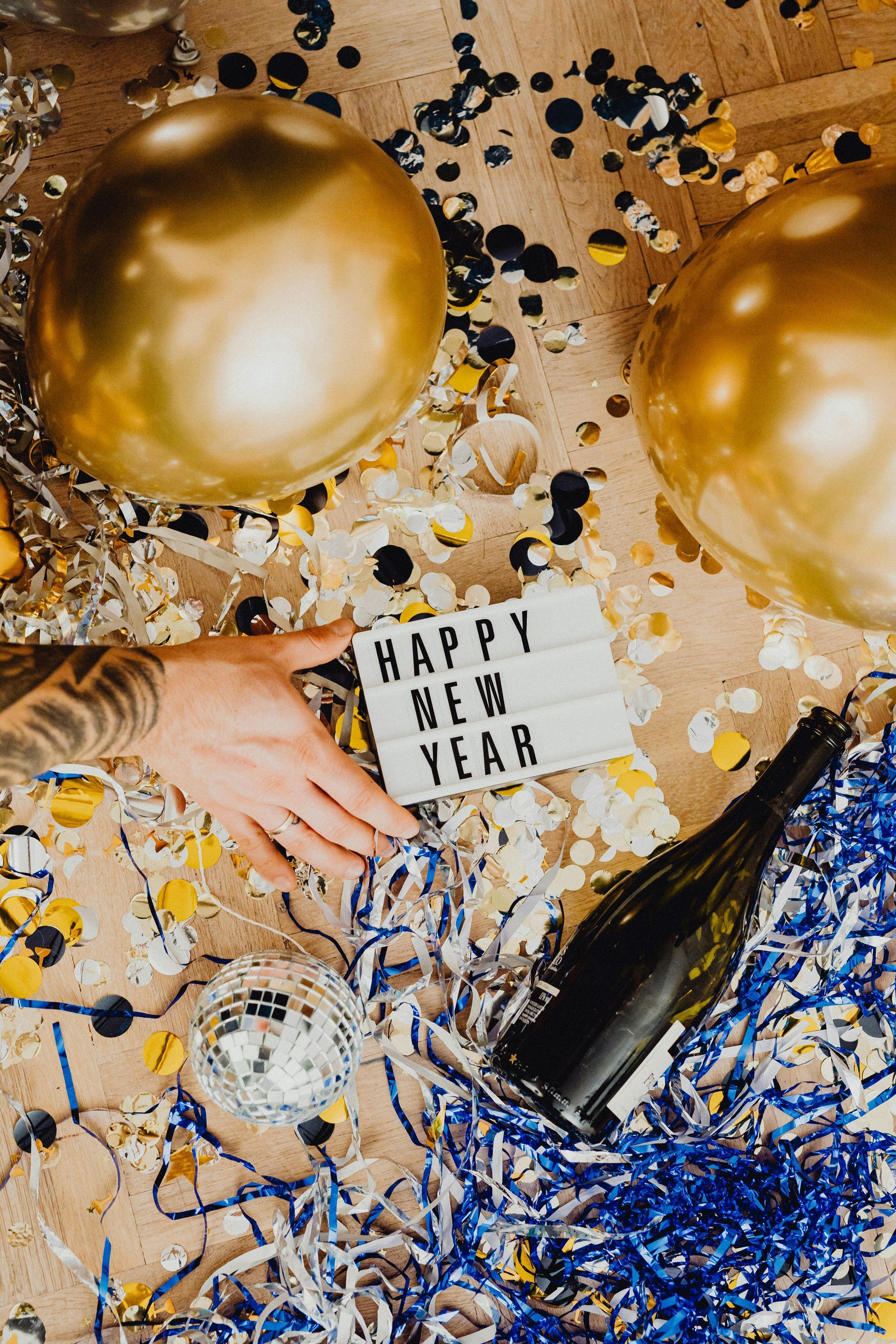 Een hand reikt naar een bordje met de tekst „Gelukkig Nieuwjaar“ te midden van gouden ballonnen, confetti en een fles champagne op de grond.