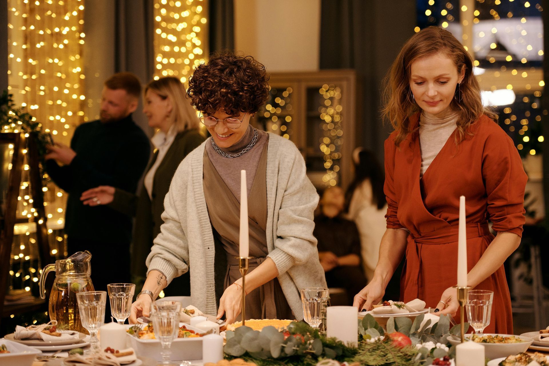 Two people are setting a festively decorated table in a room warmly lit by string lights and getting ready for a cozy get-together.