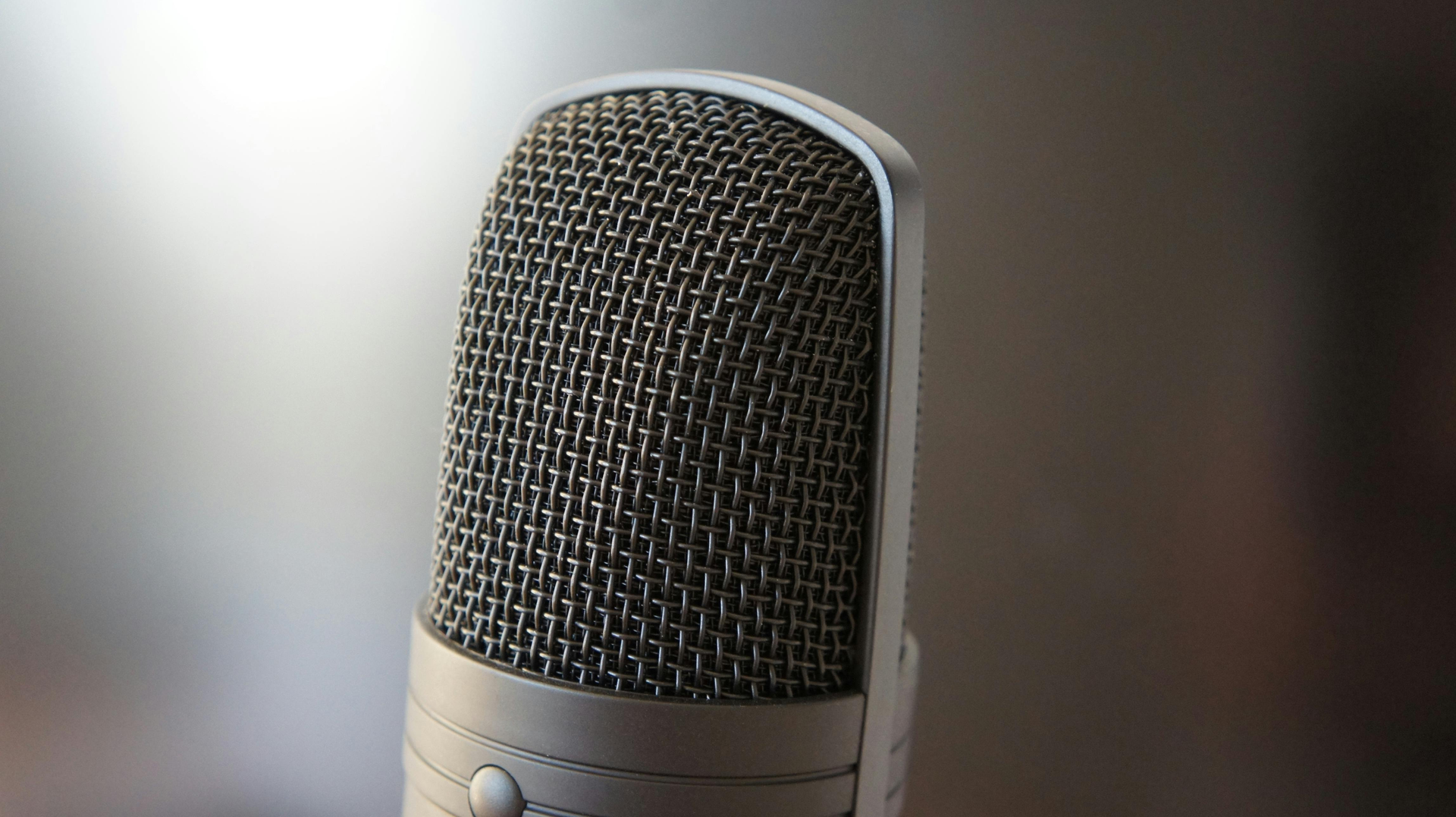 A close-up of a silver studio condenser microphone covered in wire mesh, set against a blurred, neutral background.